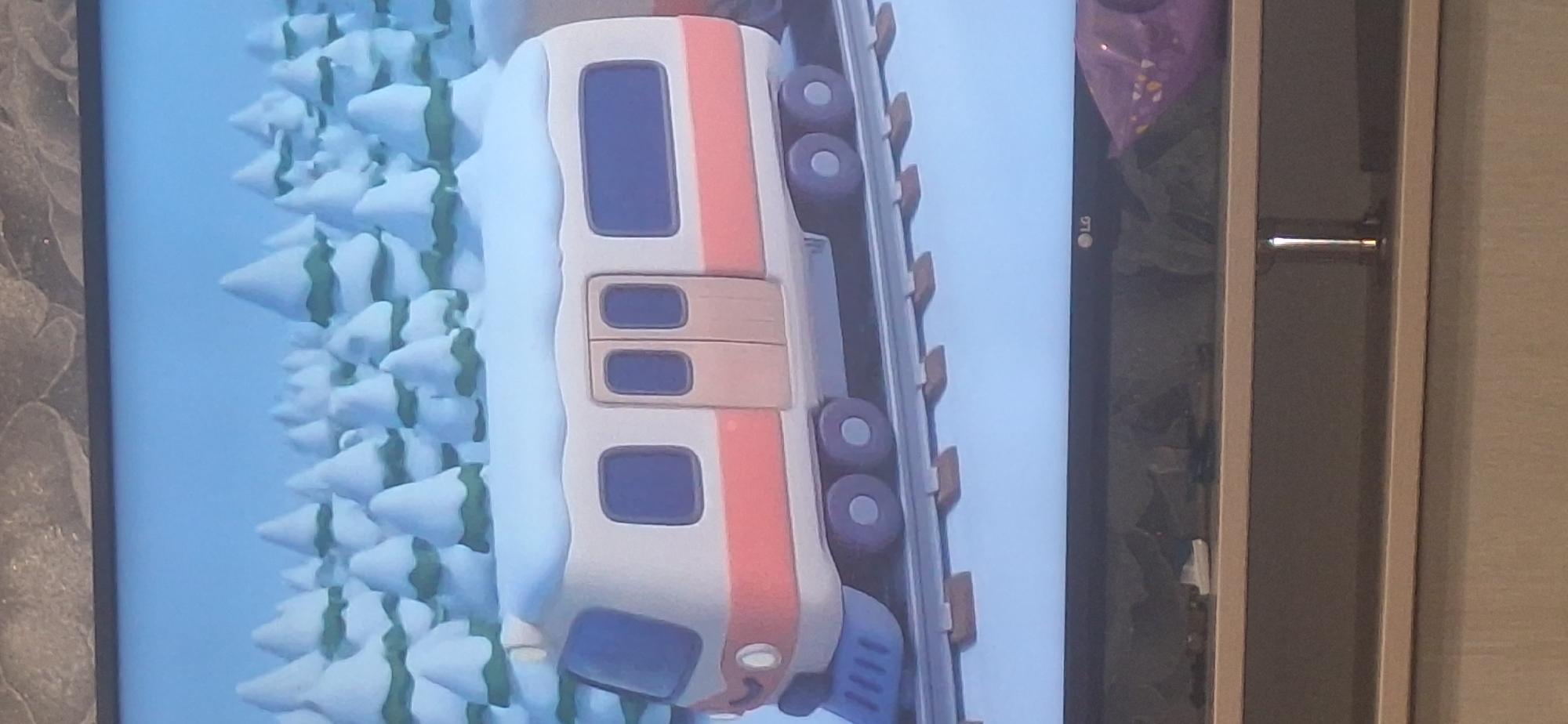 The image shows a cartoon of a train in a snowy forest. The train is white with a pink stripe and has blue windows. It is on a track that is covered in snow. In the background, there are many snow-covered trees. The image is of a television screen, with the bottom part of the screen showing a shelf and a portion of the floor. The LG logo is visible on the bottom edge of the television screen. No specific location in Bila Tserkva, Ukraine is discernible from the image.