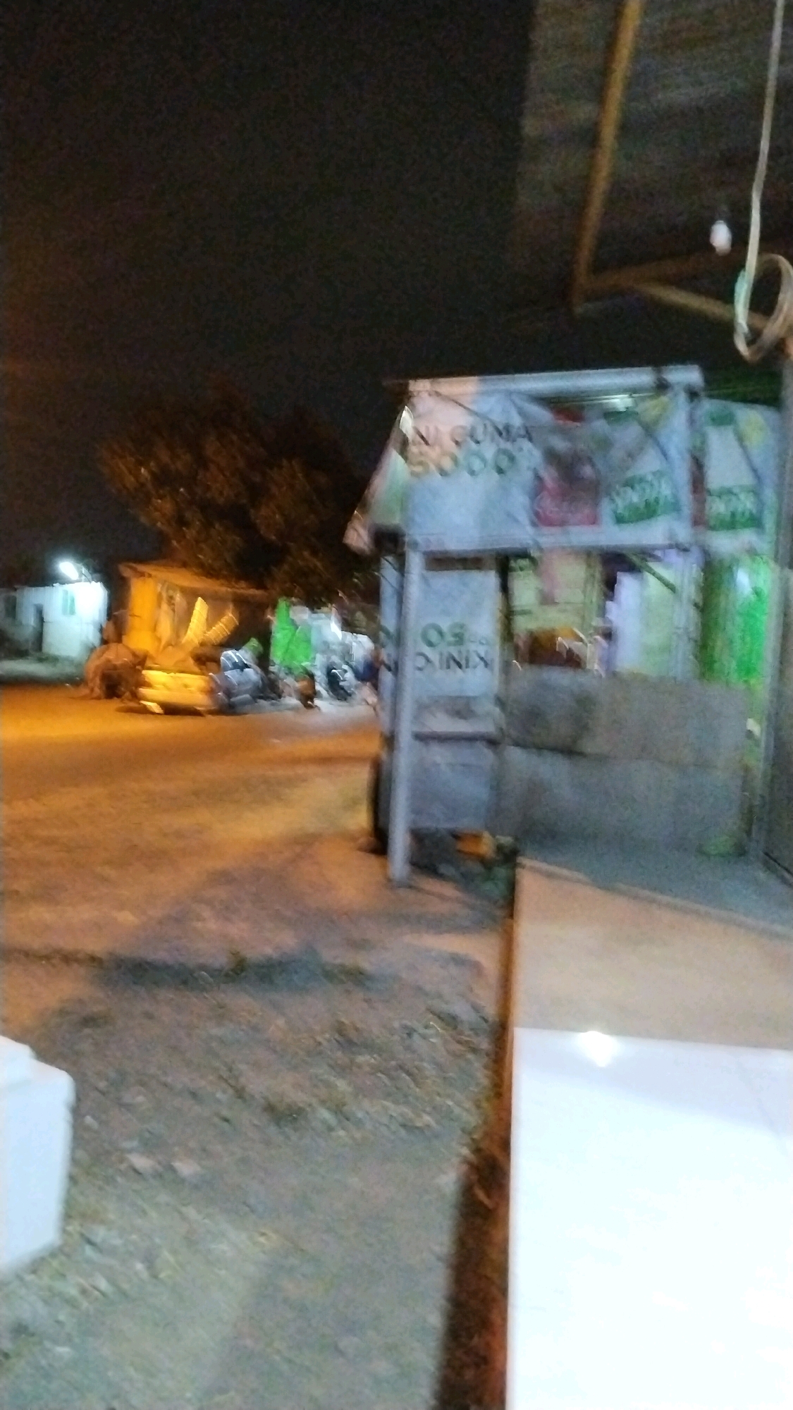 A blurred, low-light photograph captures an outdoor scene in Balongmojo, Indonesia, likely at night. In the foreground, a rough, uneven ground surface is visible. To the right, a portion of a lighter-colored structure, possibly a building wall or counter, occupies the edge of the frame. Dominating the mid-ground is a small, open-air stall or kiosk with a light-colored, possibly plastic, roof supported by a metal frame. Various items are displayed or stored around and within the stall, including what appear to be bags of goods stacked behind the counter. Some of these bags have text on them, with prominent green and white lettering visible. A sign on the front of the stall reads "KING OS 50". To the left of the stall, a pile of what looks like bundled wood or construction materials is present. Further in the background, a building with lights on can be seen, along with trees. The overall impression is a somewhat cluttered street scene at dusk or after dark.