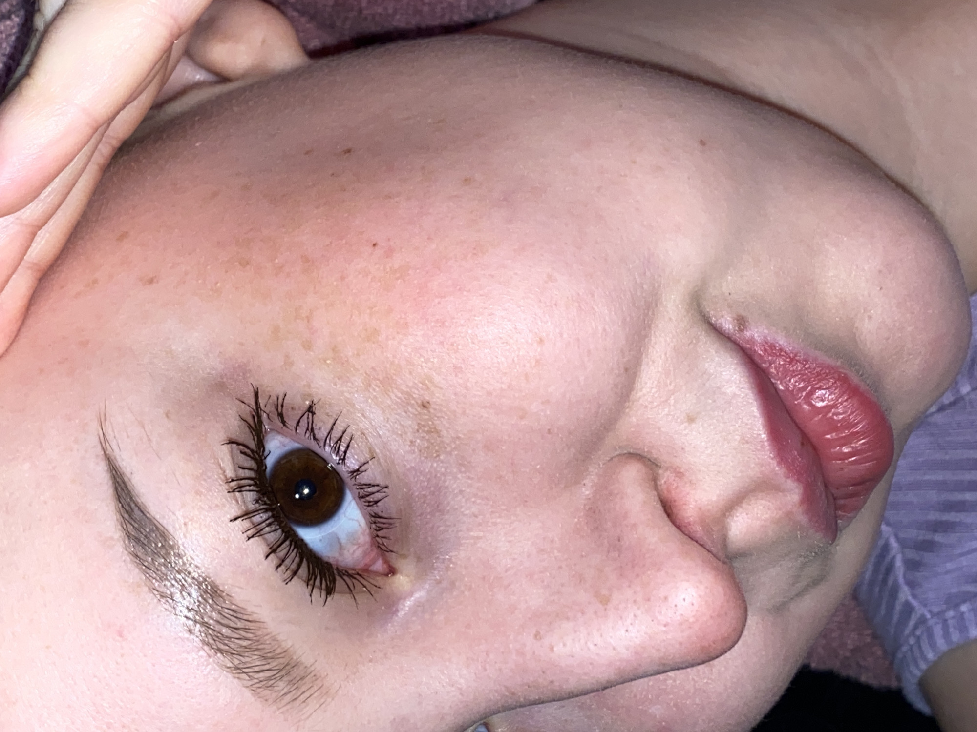 The image is a close-up of a person's face, tilted to the side. The person has fair skin with freckles. Only one eye is visible, brown in color, with prominent eyelashes coated in mascara. An eyebrow is visible above the eye, appearing groomed. The nose and lips are also visible. The lips are colored with a red lipstick. The environment is blurry and undefined, making it difficult to discern the location in Chaikovsky, Russia. No specific actions or interactions are observable beyond the presentation of the face.