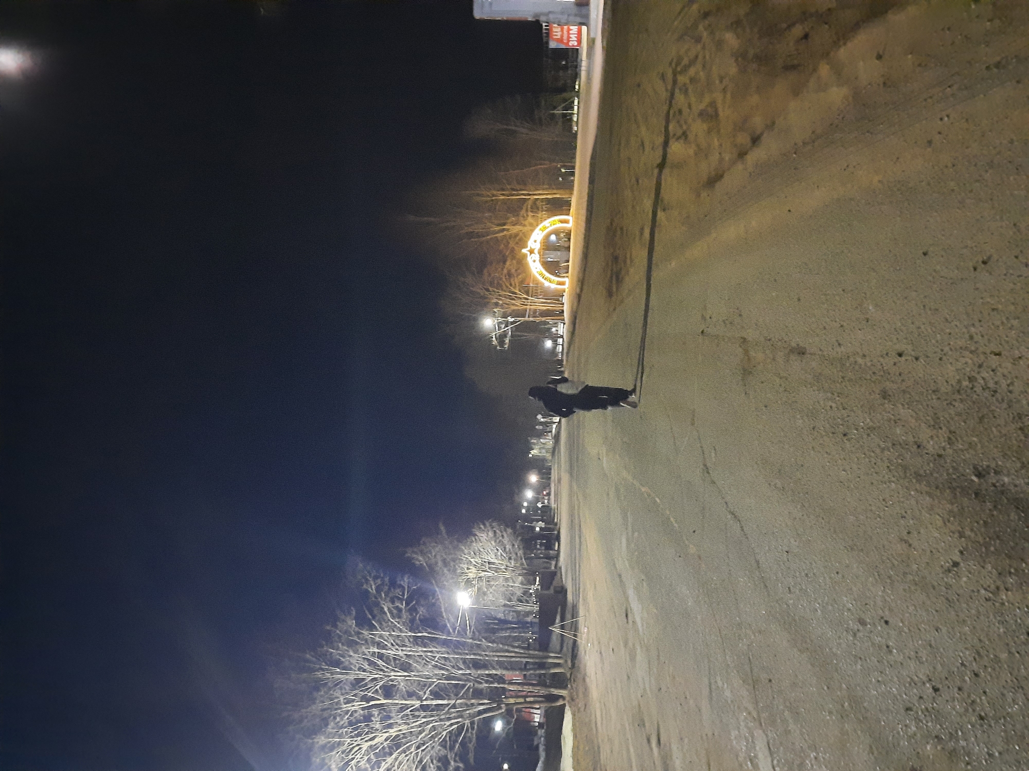 A nighttime view of a street in Krasnogorskoye, Russia. The pavement is visible and slopes upwards from left to right. A figure is seen on the right side of the road. Streetlights illuminate the scene, and bare trees line the left side. In the distance, there is an illuminated decorative arch. A building with Russian signage is visible in the far right background. The sky is dark.