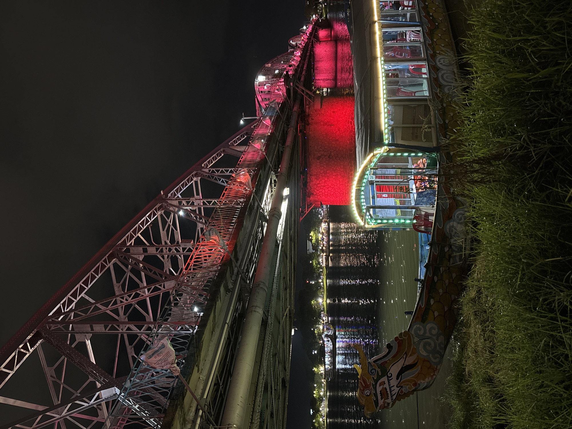 This is a night photograph taken in Phú Xuân Ward, Vietnam, capturing a dragon boat docked by the river, with a bridge illuminated in red overhead. The dragon boat, adorned with colorful traditional patterns, rests on a grassy bank. Its interior is lit with yellow string lights, revealing red chairs and other passengers inside. The river's surface reflects the lights from the bridge and distant buildings, creating a vibrant, shimmering effect. The bridge, a prominent structure, features red lighting along its railings and arches, with its stone pillars also bathed in a strong red glow. The background shows a tree-lined promenade with more lights, indicating a populated urban area along the river. The sky is dark and cloudy, characteristic of nighttime.