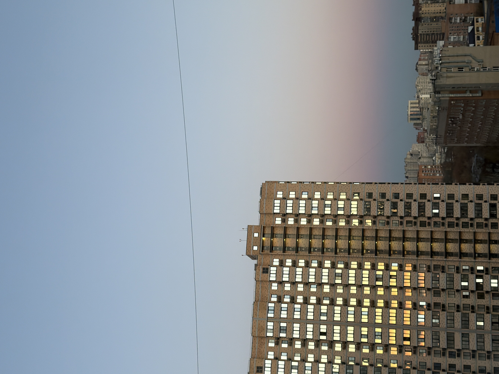 The image depicts a partially visible high-rise building in Rostov-on-Don, Russia, at dusk or dawn. The building, constructed of concrete or stone, features a grid-like façade with numerous windows. Many of the windows are illuminated, suggesting the presence of inhabitants. In the background, the silhouettes of other buildings are visible against a sky transitioning from blue to a light pinkish-grey. A single power line is visible spanning across the sky. The scene is captured from a low angle, emphasizing the height of the building.
