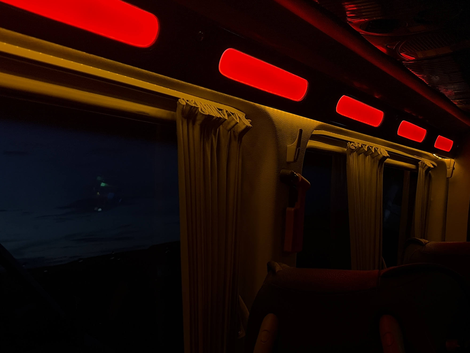 The interior of a train car at night. Red rectangular lights illuminate the ceiling. Tan curtains hang over the windows. Outside the window, a dark landscape is visible. The background is completely black. Seats are in the foreground. The location of Anipemza, Armenia, is not discernible from the image.