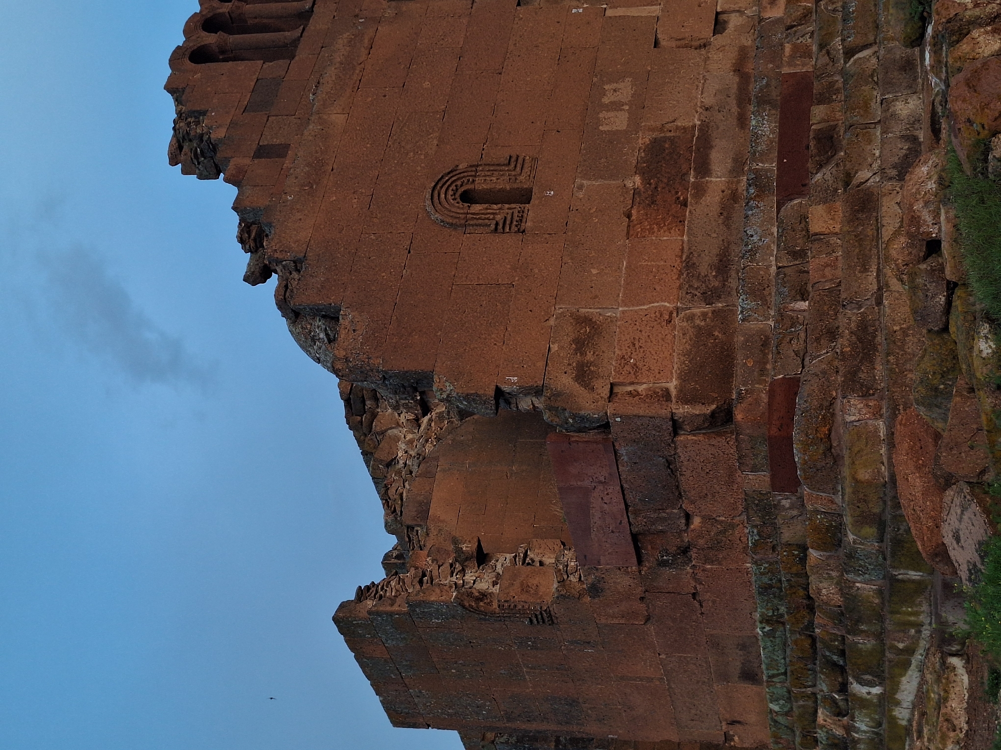 The image shows a section of a ruined building in Anipemza, Armenia. The building is constructed of red brick. The wall is in a state of disrepair, with significant portions of the brickwork missing. There is a decorative carving resembling a "U" shape. The background shows a blue sky.