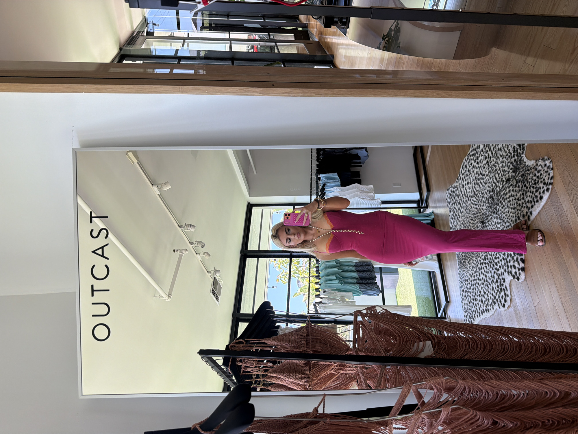 A vertical shot of a woman in a mirror, standing inside a clothing store in West Hollywood, California. The reflection shows the blonde-haired woman is wearing a hot pink dress and holding a pink cell phone, presumably taking a selfie. The backdrop features clothing racks with various items and the store's signage "OUTCAST." The floor is made of hardwood, and there is a black and white patterned rug in the background. Outside the window, green vegetation is visible.