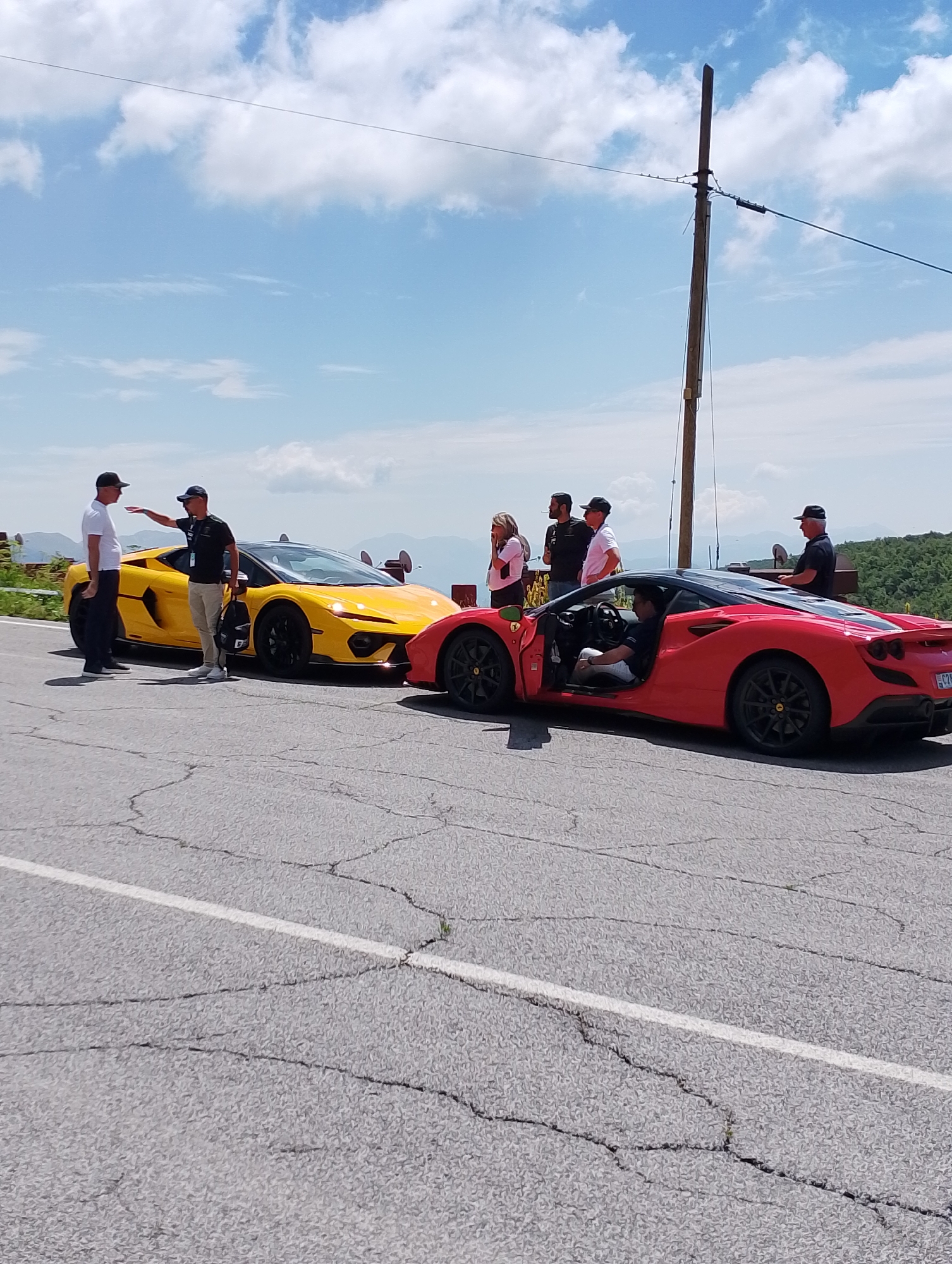 In Micigliano, Italy, on a clear day, a group of people are gathered next to two high-performance sports cars parked on a cracked asphalt road. The vehicles include a bright yellow Lamborghini and a red Ferrari, both featuring sleek, aerodynamic designs and black accents. Several individuals are visible, some interacting with each other and the cars, while others observe. One man in a white shirt and black cap is seen talking to another man in a dark shirt and cap, with the yellow Lamborghini positioned between them. The red Ferrari's driver's side door is open, and a person is seated inside. In the background, a utility pole with wires stands tall against a sky filled with fluffy white clouds. Rolling hills covered in lush green foliage extend into the distance, suggesting a scenic, possibly mountainous, location. The overall scene captures a moment of leisure and appreciation for luxury automobiles in a picturesque Italian setting.