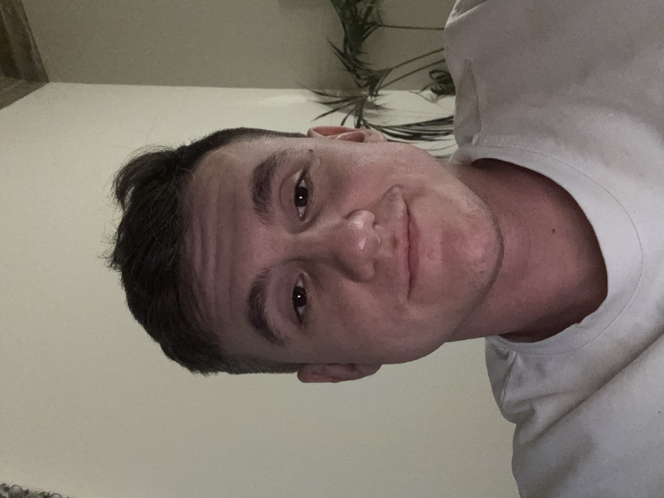 A male subject is depicted in an indoor environment, seen from the chest up in a close-cropped frame. He has short, dark hair, a light complexion, and is looking directly at the camera with a slight smile. He is wearing a plain white crew-neck t-shirt. The background consists primarily of a light-colored, undecorated wall. In the upper right corner, a portion of a textured ceiling or beam structure is visible. To the right of the subject's head, the dark green leaves and stems of an indoor plant are partially visible against the wall. No visual elements within the image provide identification of a specific city or location as Venice, Italy.