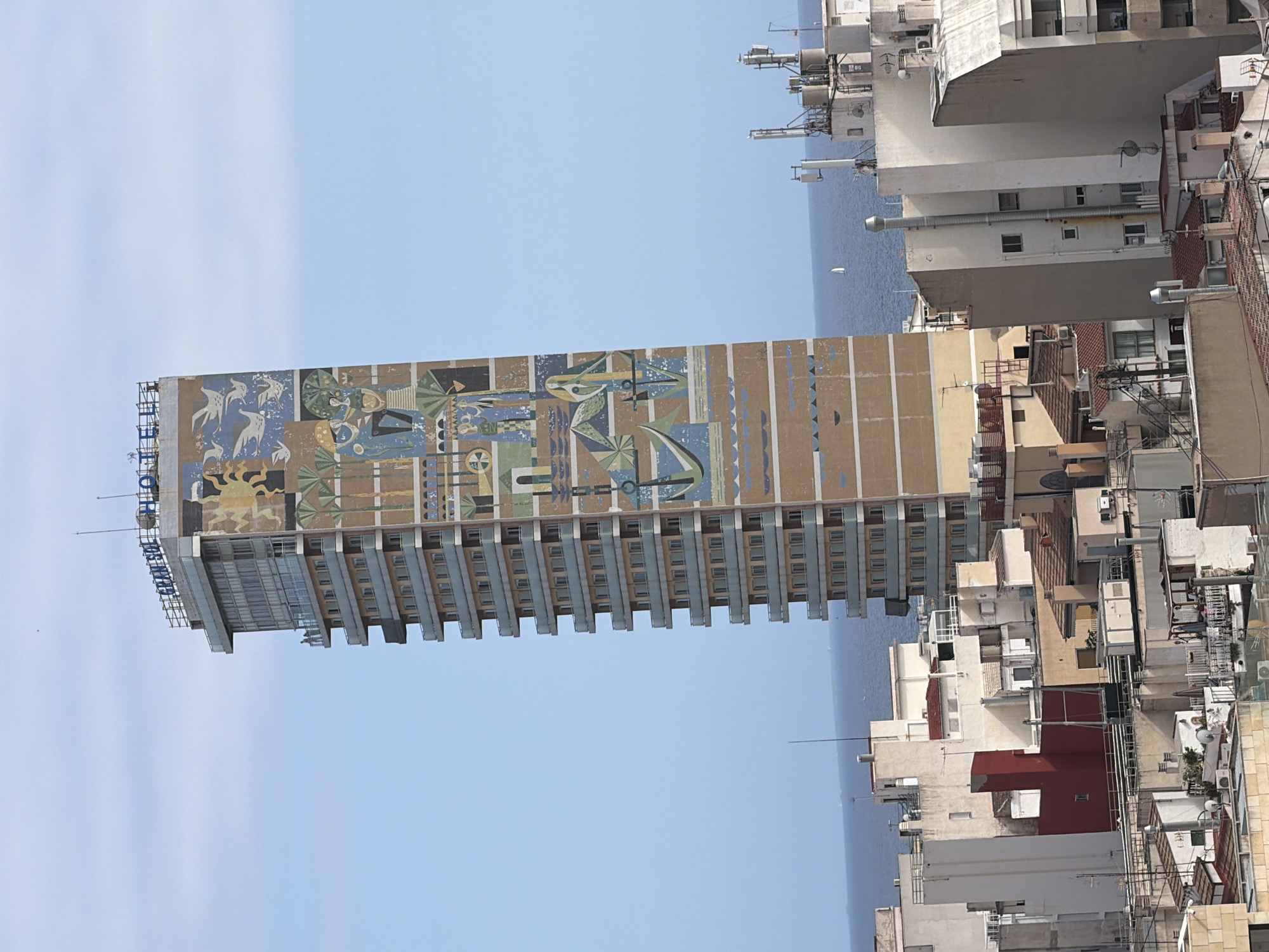 An eye-level, slightly high-angle, full shot captures the facade of a tall, rectangular building with a mural on its north-facing side, set against a pale blue sky and a dark blue ocean. The building, which appears to be a hotel, has a name sign reading "GRAN SOL HOTEL" in blue block letters at its apex. The mural, composed of geometric shapes and stylized figures, depicts a scene with various elements including palm trees, birds in flight, human figures, and a large anchor motif. The building has numerous balconies with glass railings, and a series of horizontal bands of windows. In the foreground and midground, other buildings of varying heights, constructed from concrete and painted in shades of white, beige, and red, form a dense urban landscape. Rooftops are a mix of flat surfaces, tiled roofs, and some with visible mechanical equipment and satellite dishes. The ocean stretches across the horizon behind the buildings, and a small white sailboat is visible in the distance. The overall impression is that of a coastal city under a clear, sunny sky.