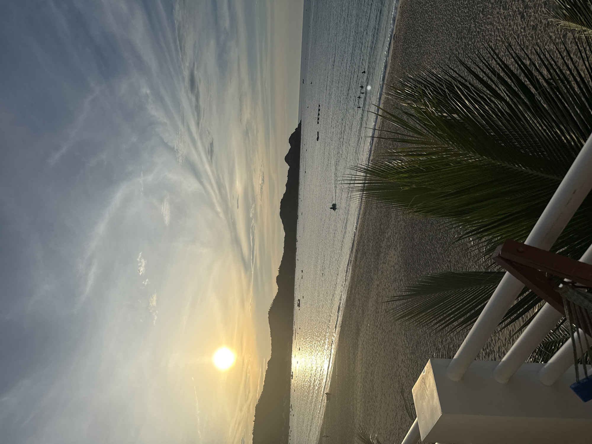 This is a photograph taken from a raised vantage point, likely a balcony or railing, overlooking a beach and the sea at sunset in Song Cau, Vietnam. The sun, a bright orb, is setting on the left side of the frame, casting a golden glow across the calm ocean and reflecting off the water in a shimmering path. In the distance, a dark, hilly coastline stretches across the horizon, silhouetted against the sky. Several small boats are scattered across the water, some closer to shore and others further out. On the beach, the sand appears light brown and gently slopes towards the water. A few figures are visible on the beach and in the shallows of the sea, suggesting recreational activity. In the foreground, the fronds of a palm tree partially obscure the view, adding a tropical element to the scene. The sky is a soft gradient of blue and white, with wispy clouds streaked across it. The overall impression is one of tranquility and the end of a day.