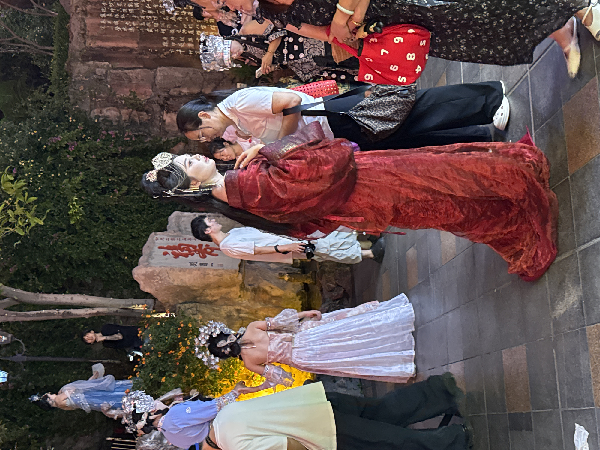 In Yuzhong District, China, a group of people are gathered outdoors at dusk. The scene appears to be a cultural performance or a themed event, given the traditional attire worn by some individuals. In the foreground, a woman in a vibrant red, floor-length traditional Chinese dress with elaborate sleeves is the central subject, facing left and looking at another woman. This second woman, wearing glasses and a white shirt, is looking at the woman in red. Behind them, a woman in a light pink traditional dress with a floral pattern and an ornate headdress stands facing away from the camera. To the left, a person in a light-colored t-shirt and dark pants is partially visible, walking away. In the background, several other individuals are present, some wearing traditional headdresses and clothing. A stone wall with Chinese inscriptions is visible behind the main subjects, and lush green foliage and trees form the backdrop. The ground is paved with square tiles. A street lamp is visible in the upper left corner, indicating an urban setting, likely illuminated for an evening event.