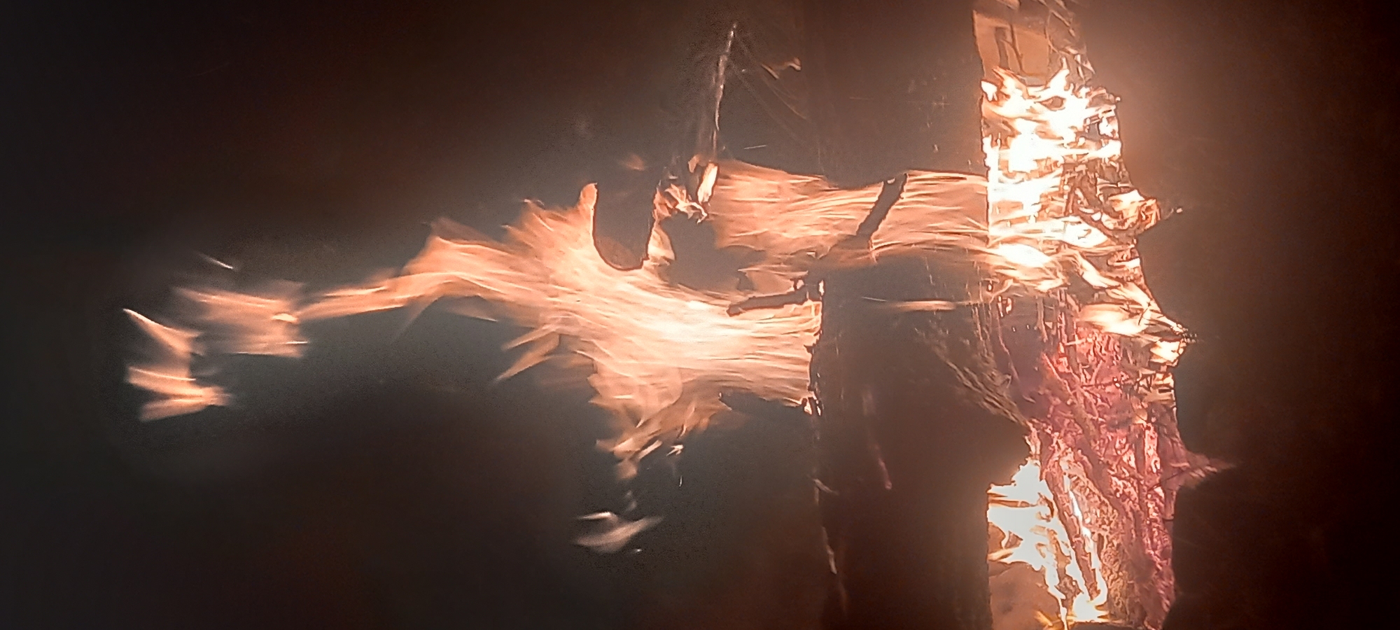 This vertical photograph captures a large, dynamic fire, likely a bonfire or pyre, burning intensely against a dark background in Kryvyi Rih, Ukraine. The flames, exhibiting hues from deep red and orange to bright yellow, surge upwards, consuming visible logs and timber arranged in multiple tiers. Smoke and embers are discernible within and around the main body of the fire. At the bottom of the frame, the dark silhouettes of several individuals' heads and shoulders are visible, positioned directly in front of the lowest flames and embers, suggesting an audience observing the event. The background is uniformly dark, providing no additional environmental context beyond the immediate vicinity of the fire, indicative of a nighttime setting.
