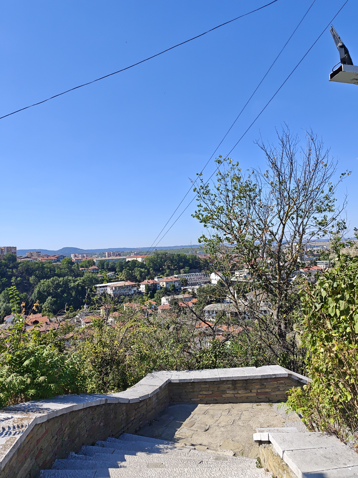 This image captures a scenic overlook in Lovech, Bulgaria, likely from the area near the Varosha quarter or the fortress. In the foreground, a stone staircase with a decorative brick and stone retaining wall leads downwards, offering a pathway through the greenery. The stairs are made of large, flat stone slabs. Beyond the immediate foreground, a lush canopy of trees and vegetation dominates the middle ground, with the town of Lovech spread out below. The houses in the town are characterized by their red-tiled roofs, a common architectural feature in the region. In the distance, rolling hills form the horizon under a clear blue sky. The scene is bathed in bright daylight, indicating fair weather. A thin black cable, possibly part of a utility line or a zipline, traverses the sky diagonally from the upper left to the upper right of the frame. A dark, metallic structure, possibly a support for the cable or part of an unseen mechanism, is visible in the upper right corner against the sky.