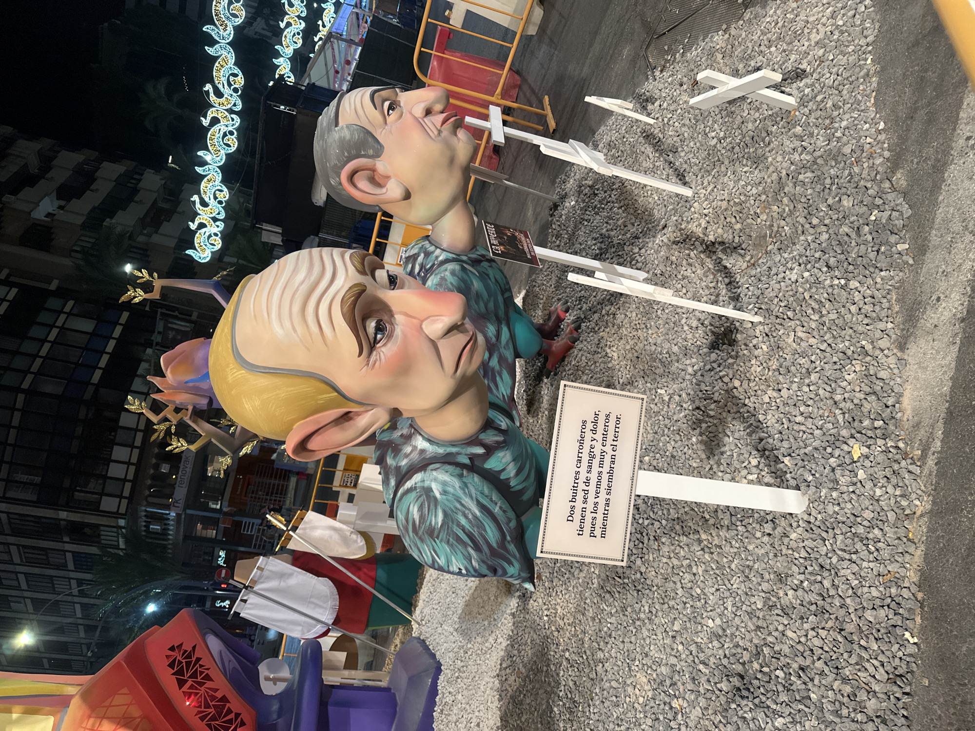 The image depicts a nighttime street scene in Alicante, Spain, likely during a festival. In the foreground, two large, caricatured heads of men are part of an artistic display. The head on the left is larger, with a blondish-yellow wig and a pained expression. The head on the right is smaller and bald, with a more stern expression. Both heads are attached to bodies that appear to be those of birds, with teal and green feathered bodies. One of the bird-like bodies is on the left and appears to be wearing a teal and green feathered outfit. The other bird-like body, on the right, is mostly obscured.
In front of the display, a sign with decorative borders reads "Dos buitres carroñeros tienen sed de sangre y dolor, pues los vemos muy enteros, mientras siembran el terror." (Two scavenger vultures are thirsty for blood and pain, for we see them very whole, while they sow terror.) White crosses, of varying sizes and some shaped like an 'X', are scattered on the gravel surrounding the display.
Behind the main display, a red and yellow barrier is visible, along with a building in the background with illuminated windows. Strings of decorative lights, a common sight during festivals, are strung across the street overhead. The ground is covered in small grey gravel. The overall impression is a political or social commentary expressed through public art during a festive event.