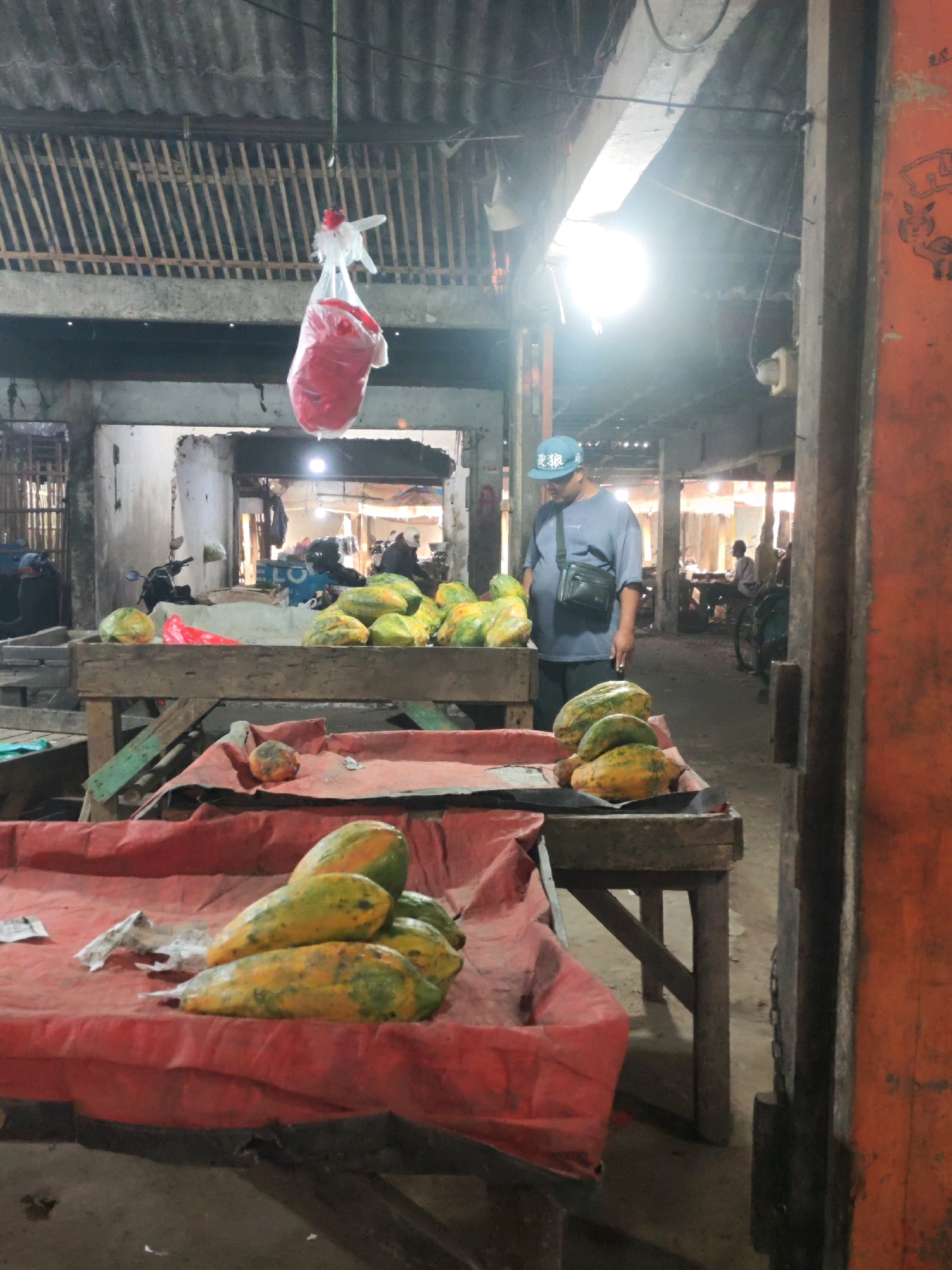 The image shows a market stall in Karawang, Indonesia. A man wearing a blue cap and a blue shirt stands behind a table piled with papayas. He is looking down at the fruit. There are other papayas on display in wooden crates and on red cloths. In the background, there are more market stalls, some with motorcycles and other vehicles. The market appears to be covered by a corrugated metal roof. A bag filled with red material is hanging from the ceiling. The overall impression is of a bustling, open-air market.