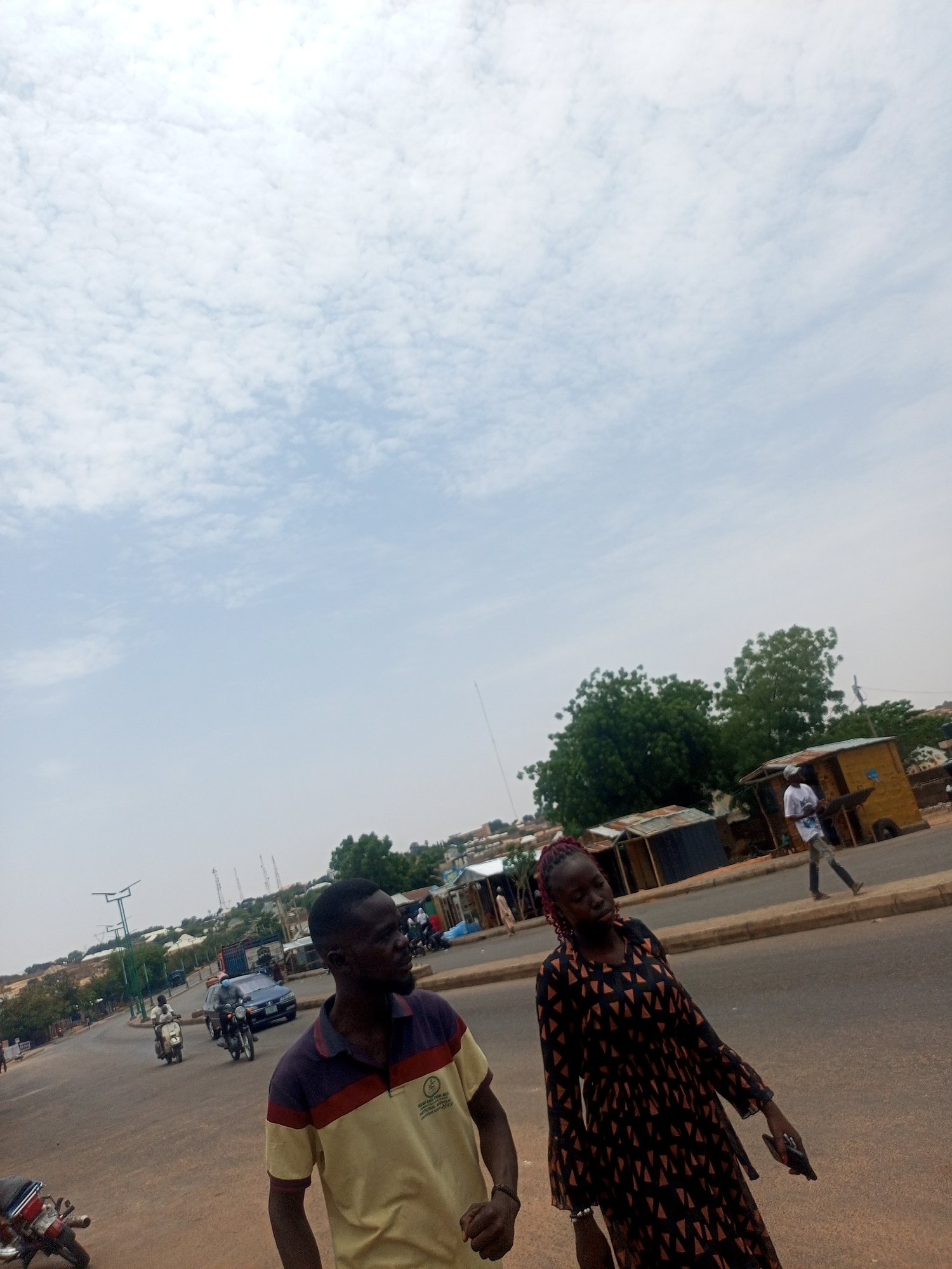 Two people, a man and a woman, are walking on a street in Tempure, Nigeria. The man is wearing a short-sleeved shirt with a red and purple stripe, and the woman is wearing a long-sleeved dress with black triangles. There is traffic on the street, including cars and motorcycles. Several simple buildings line the side of the road, beyond a curb. The sky is bright with scattered clouds.