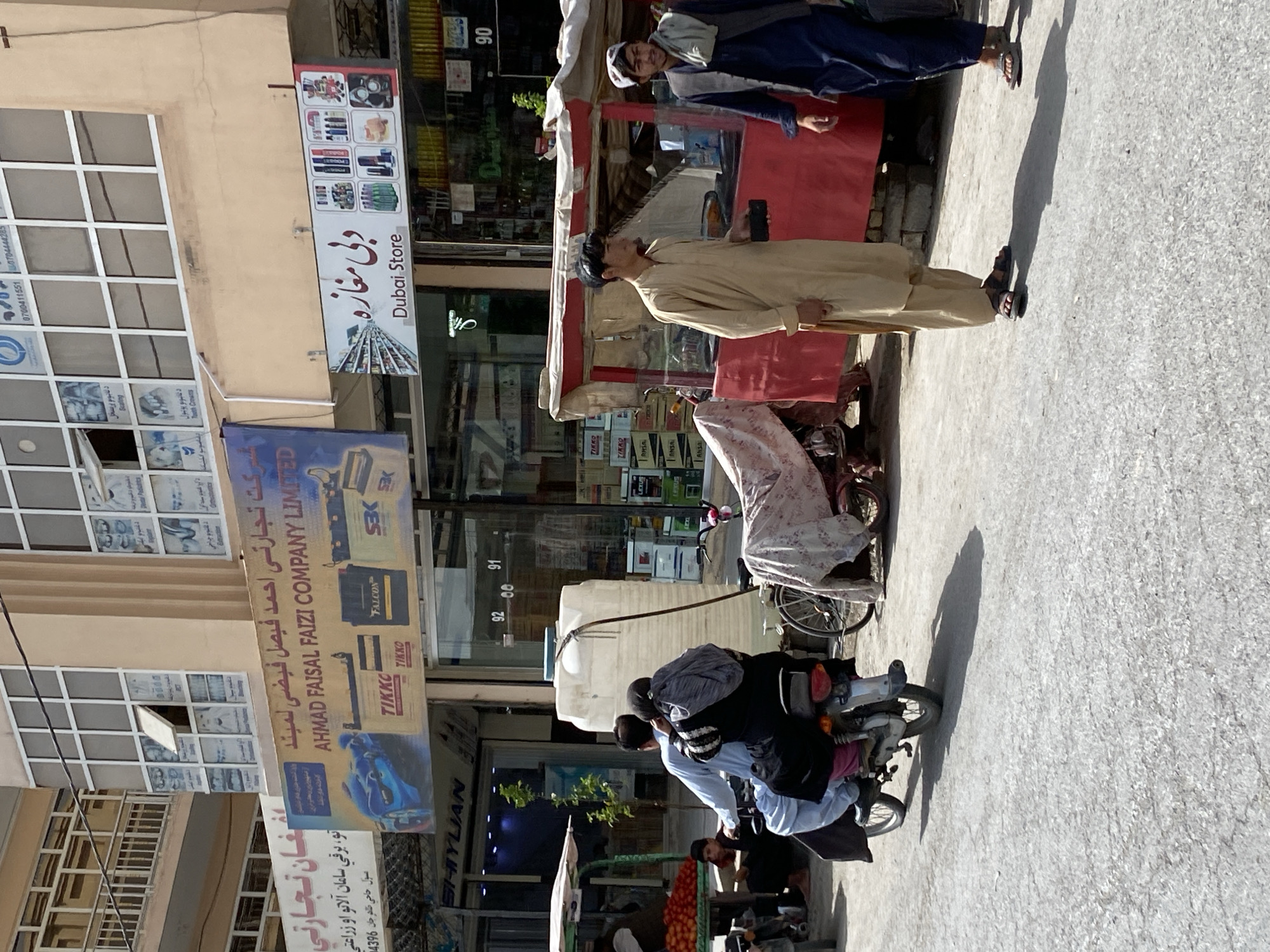 This image depicts a street scene in Kandahar, Afghanistan. In the foreground, a man and a woman are riding on a motorcycle. The man is driving, and the woman is sitting behind him, holding onto him. Both are dressed in traditional Afghan clothing. The motorcycle is silver and has a large white water tank attached to the back. In the background, there are several shops and stalls. One shop has a sign that reads "AHMAD FAISAL FAIZI COMPANY LIMITED" and displays images of car batteries. Another shop has a sign that says "Dubai Store". There are also people walking and standing around the stalls. The overall atmosphere is that of a busy marketplace.