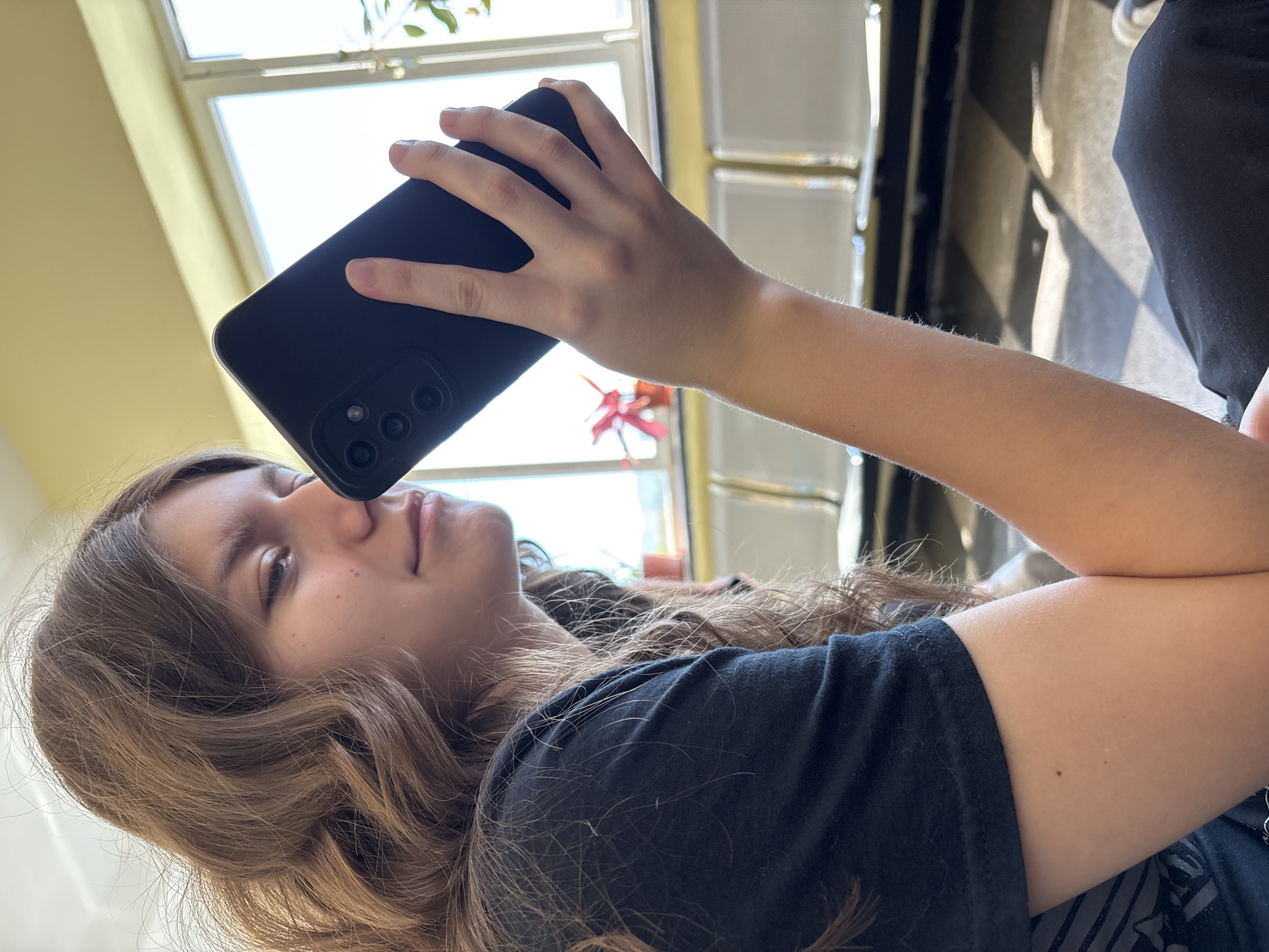 A young woman is holding a black cell phone near her face, seemingly using it to take a picture. She has brown, wavy hair and is wearing a black t-shirt. The background shows a window with plants, and a checkered floor. The location appears to be indoors. The city of Panagyurishte, Bulgaria, cannot be determined from the visual data.