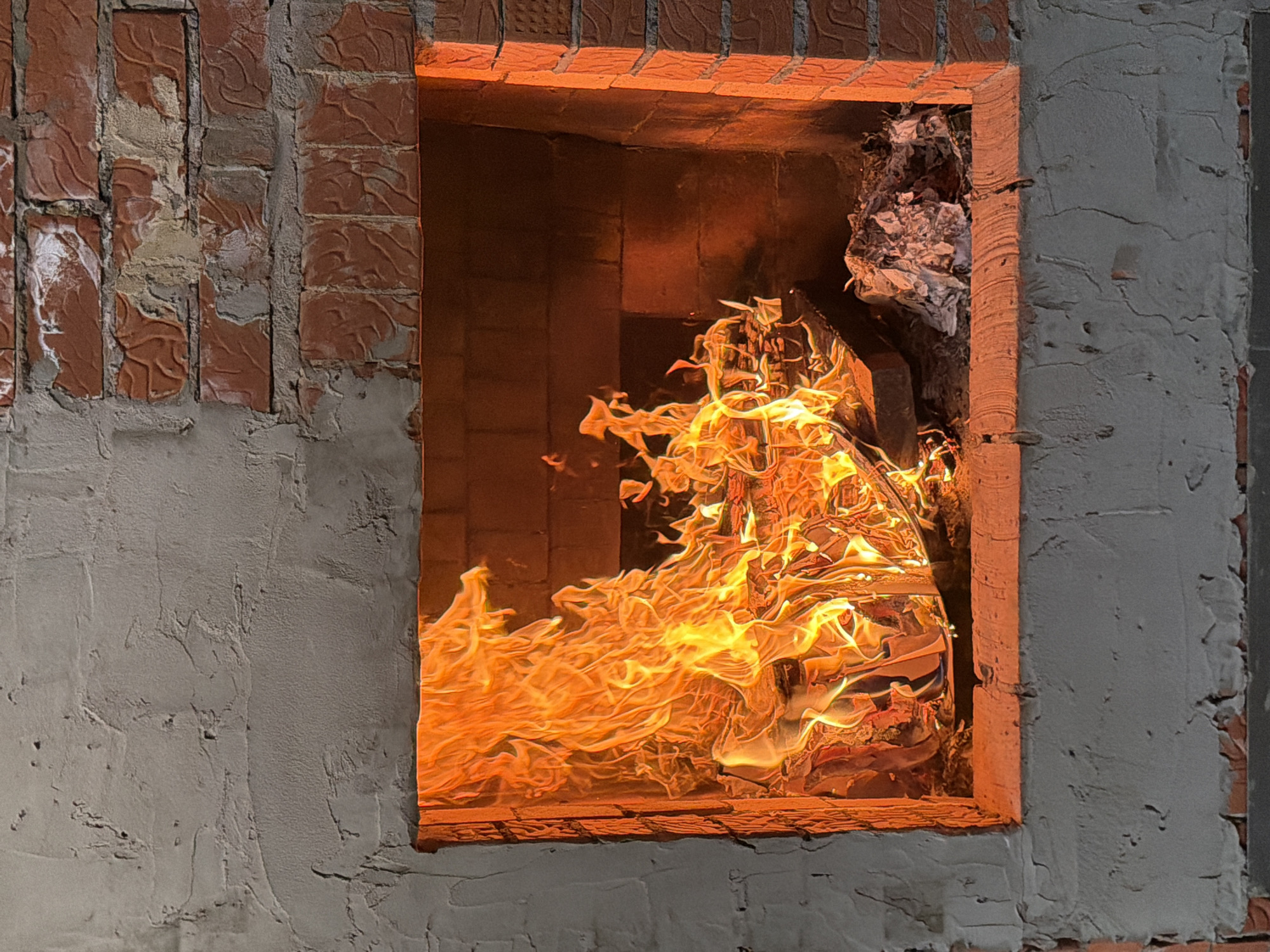A rectangular opening framed by brick and mortar is filled with a roaring fire. The fire's flames are predominantly orange and yellow, swirling and consuming what appears to be crumpled paper or similar material. The brickwork around the opening appears unfinished, with some areas covered in grey mortar. The scene suggests a furnace or a makeshift incinerator. There is no indication of the location.