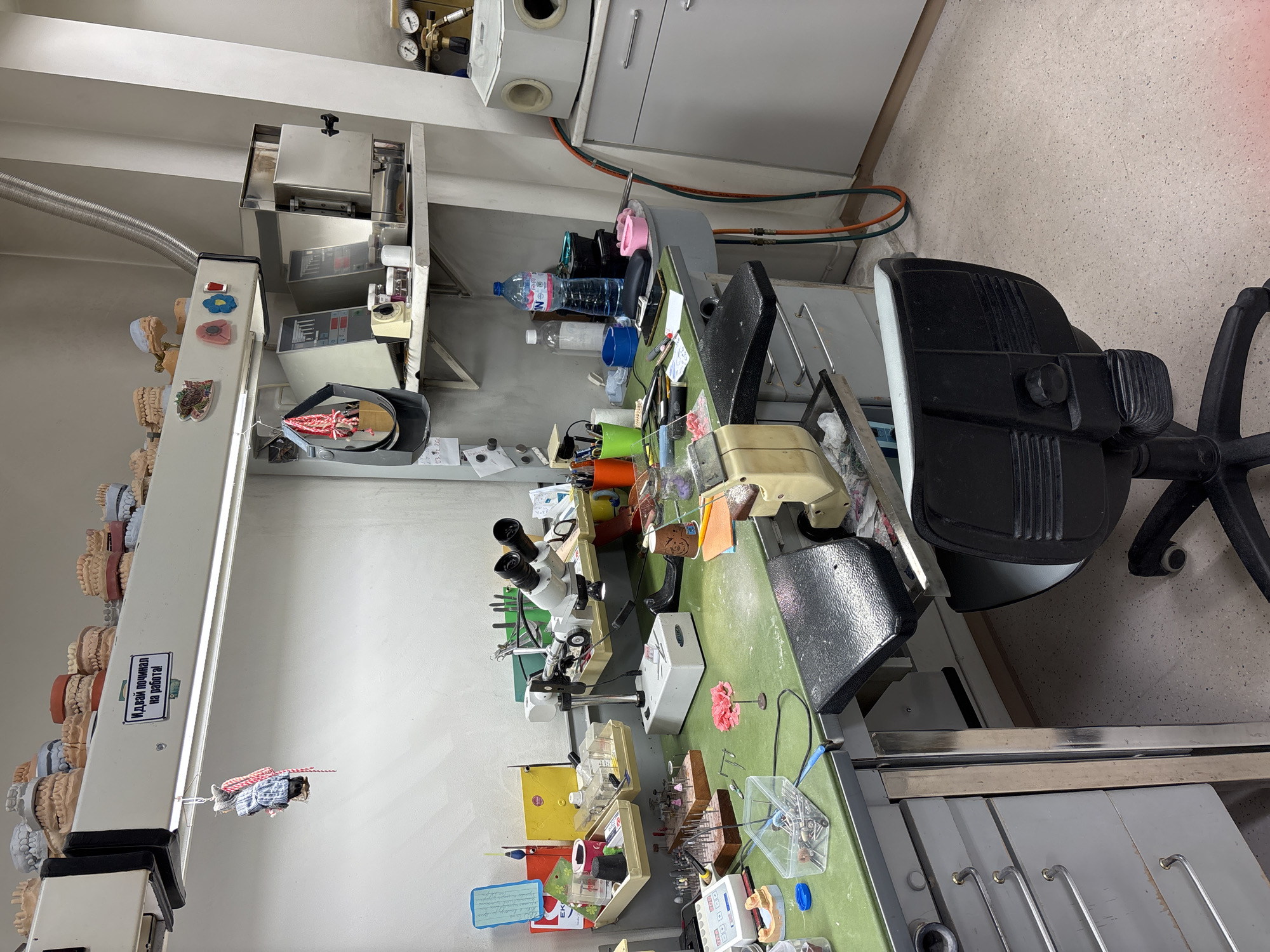 The image shows the interior of a dental laboratory, likely located in Sofia, Bulgaria, judging by the Cyrillic text on a sign. The focal point is a cluttered workbench, covered in tools and materials associated with dental prosthetics and dental repair. A microscope is prominent on the bench, along with various containers, models of teeth and jaws, hand tools, and a polishing lathe. The bench itself is a light green color and has metal drawers built underneath. A black office chair is positioned next to the bench, suggesting it is a work area. On a shelf above, there is additional equipment, including a furnace or oven and containers of liquids, including water bottles. Dental casts are arranged on a vertical fixture on the left side of the image. The room is well-lit with overhead fluorescent lighting. The scene appears to be a professional workspace used for the fabrication and repair of dental prosthetics.