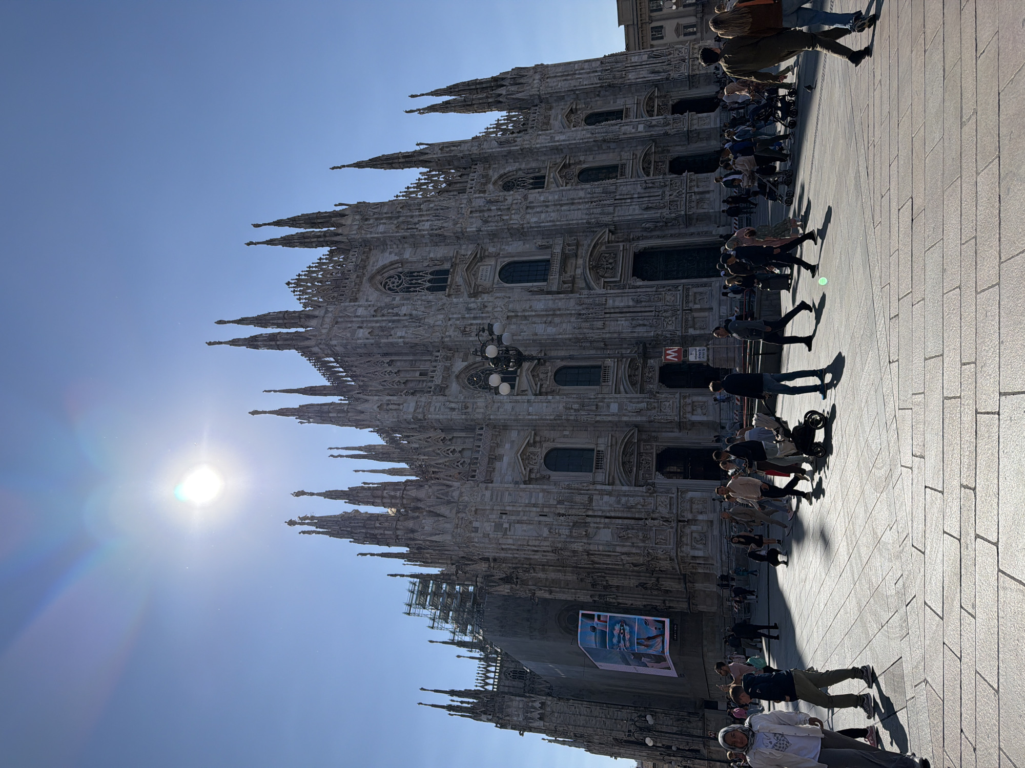 This image captures a vibrant scene in front of the Milan Cathedral (Duomo di Milano) in Milan, Italy, under a bright, clear sky.
The majestic Gothic cathedral dominates the background, showcasing its intricate white marble facade adorned with countless spires, statues, and elaborate architectural details. Many arched windows and rose windows are visible across its massive structure. A section of the cathedral on the lower left appears to have scaffolding, indicating ongoing maintenance or restoration work. Attached to this scaffolding is a large, illuminated digital display or banner featuring colorful artwork or an advertisement, though specific details are indistinct.
In the foreground and mid-ground, a paved pedestrian square extends across the scene. Numerous people are present, walking and strolling in various directions across the sunlit piazza. Their figures cast long, distinct shadows towards the viewer's right, suggesting it is either late morning or early afternoon. The ground is made of large, light-colored square tiles.
The weather is bright and sunny, with a deep blue, cloudless sky. The sun itself is prominently visible in the upper left portion of the image, causing a strong lens flare and a subtle rainbow effect.
Notable objects include a multi-globular street lamp positioned in the lower-middle and a red "M" sign, characteristic of the Milan Metro, affixed to a pole. People are dressed in casual attire, and some push strollers, indicating a leisurely atmosphere. No specific event is unfolding beyond the general activity of people congregating and passing through a popular public space.