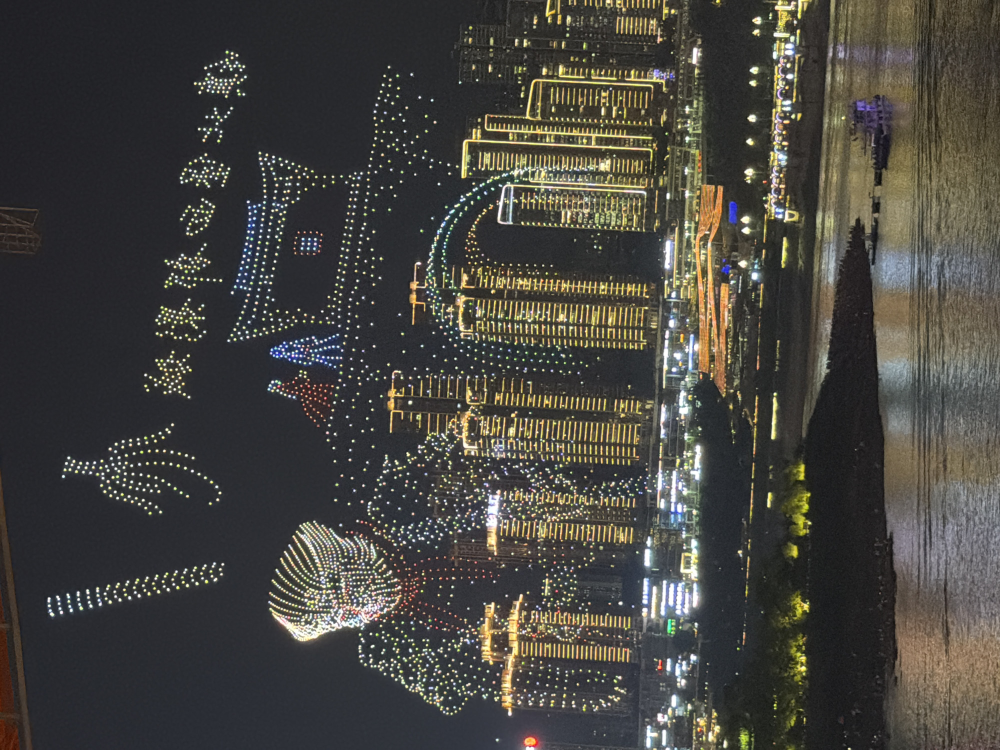 This is a nighttime aerial photograph of a city skyline and a body of water, likely a river. The scene is dominated by numerous illuminated objects and structures.
In the foreground, a drone light show is taking place, forming various shapes and patterns in the dark sky. These lights are predominantly white and yellow, with some scattered red, blue, and green lights. We can discern shapes that resemble text, a bird or dove, and abstract geometric patterns. A significant formation appears to be a stylized building or pagoda.
The background features a dense cityscape with many illuminated buildings. These structures display a multitude of windows and architectural lighting, predominantly in yellow and white. The city appears to be built along the waterfront.
To the right of the city, a large body of water reflects the city lights, creating streaks of light on its surface. A dark, triangular landmass, possibly a pier or a natural rock formation, juts out into the water from the lower right corner. A small, dark vessel with purple lights is visible on the water.
The time of day is clearly night, and the weather appears clear, allowing for the drone show and the visibility of the cityscape. The location is identified as Yuzhong District, China, which is known for its riverside urban landscape. The overall impression is one of a vibrant, modern city celebrating with a spectacular light display. There is no clear indication of people being directly visible, but their presence is implied by the organized nature of the drone show and the populated cityscape.