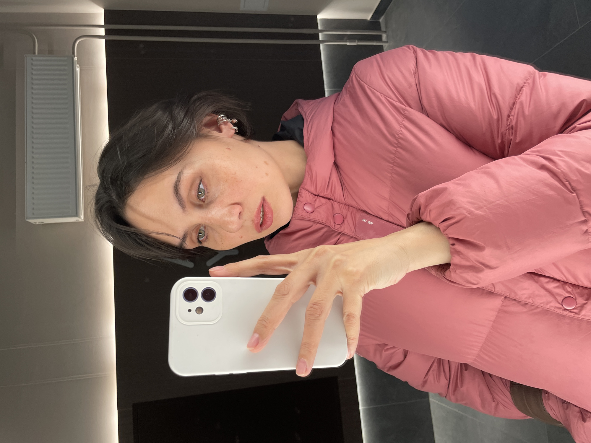 The image is a mirror selfie taken by a young person, likely a woman, in an indoor setting. She has short dark hair, light-colored eyes, and noticeable freckles across her nose and cheeks. Her expression is neutral, and she looks directly towards the phone's camera reflection.
She is wearing a dark pink, puffy winter jacket. In her right hand, she holds a white smartphone, which appears to be an iPhone, judging by the design of its dual-lens camera module. The phone is positioned to capture her reflection in the mirror.
The background suggests a modern interior, possibly a changing room or a well-lit hallway, consistent with an urban setting like Izhevsk, Russia. The walls are dark, appearing to be tiled or paneled. On the far left, a light-colored, vertically ribbed object, possibly a radiator or a ventilation grill, is visible. A vertical strip of bright artificial light runs along the right side of the frame, indicating either an illuminated mirror edge or a prominent light fixture. On the lower right side of her jacket, some visible text can be seen, which reads "АНГ. ОЗН". The lighting is artificial and bright, typical of indoor commercial spaces.