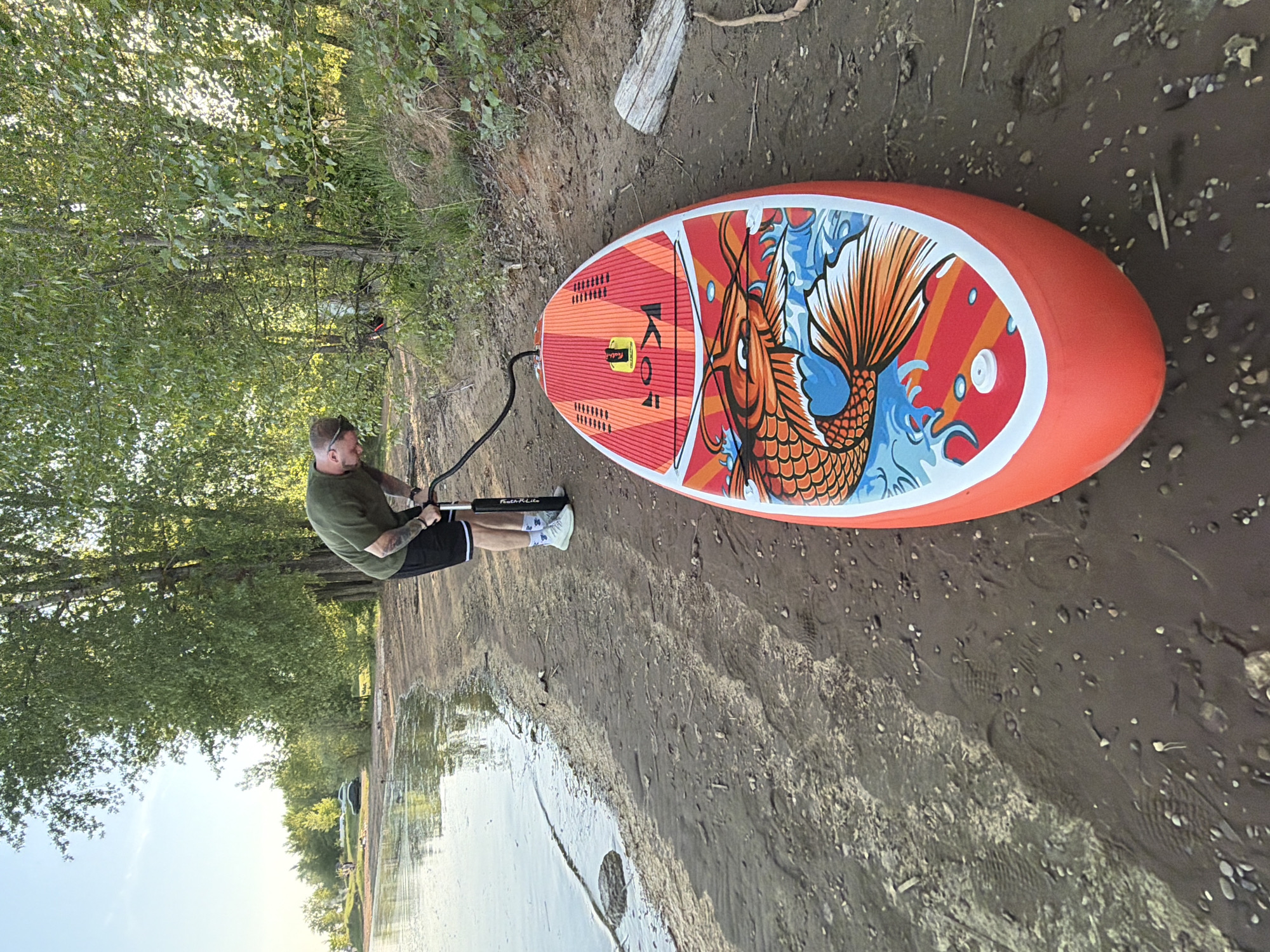 The image depicts a man inflating a stand-up paddleboard (SUP) on a muddy shoreline. The paddleboard is bright orange with a large graphic of a koi fish. The man is wearing a green shirt, black shorts with a white stripe, and white sneakers. He is holding a black pump connected to the paddleboard. The background includes trees and water, indicating a lakeside or riverside location. The scene appears to be set in a natural or park-like environment. There is no information to indicate that it is Noviy, Russia.