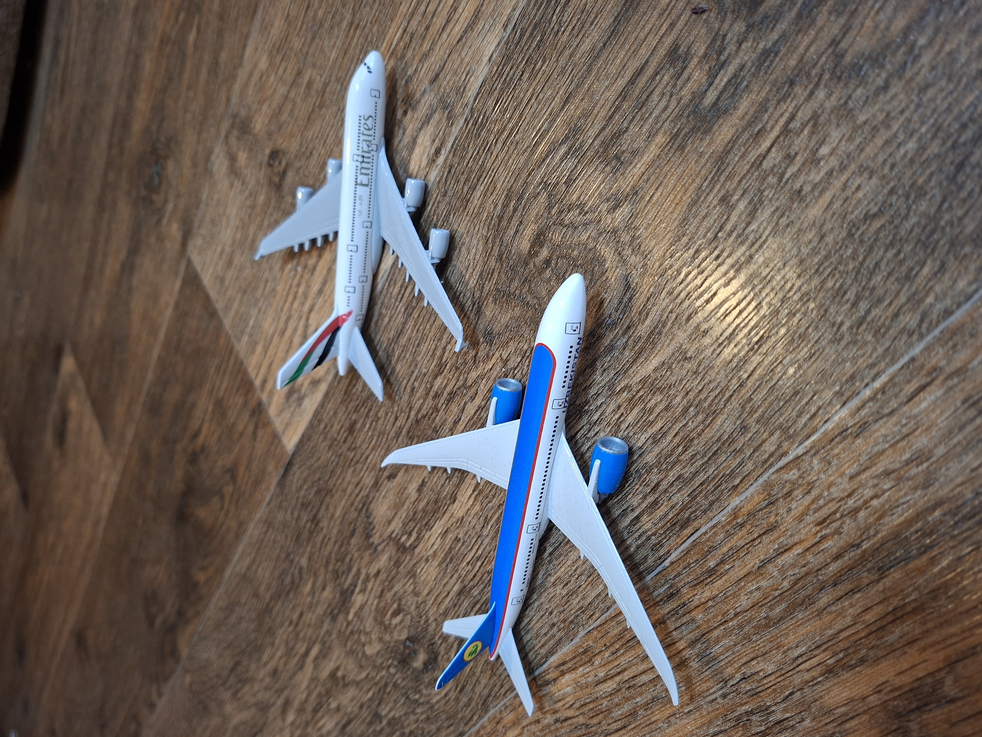 Two model airplanes are on a wooden floor. The airplane in the foreground has a blue stripe along its side and the logo of Uzbekistan Airways on its tail. The airplane in the background is white with the Emirates logo on its side. The floor is made of wooden planks with a visible wood grain.