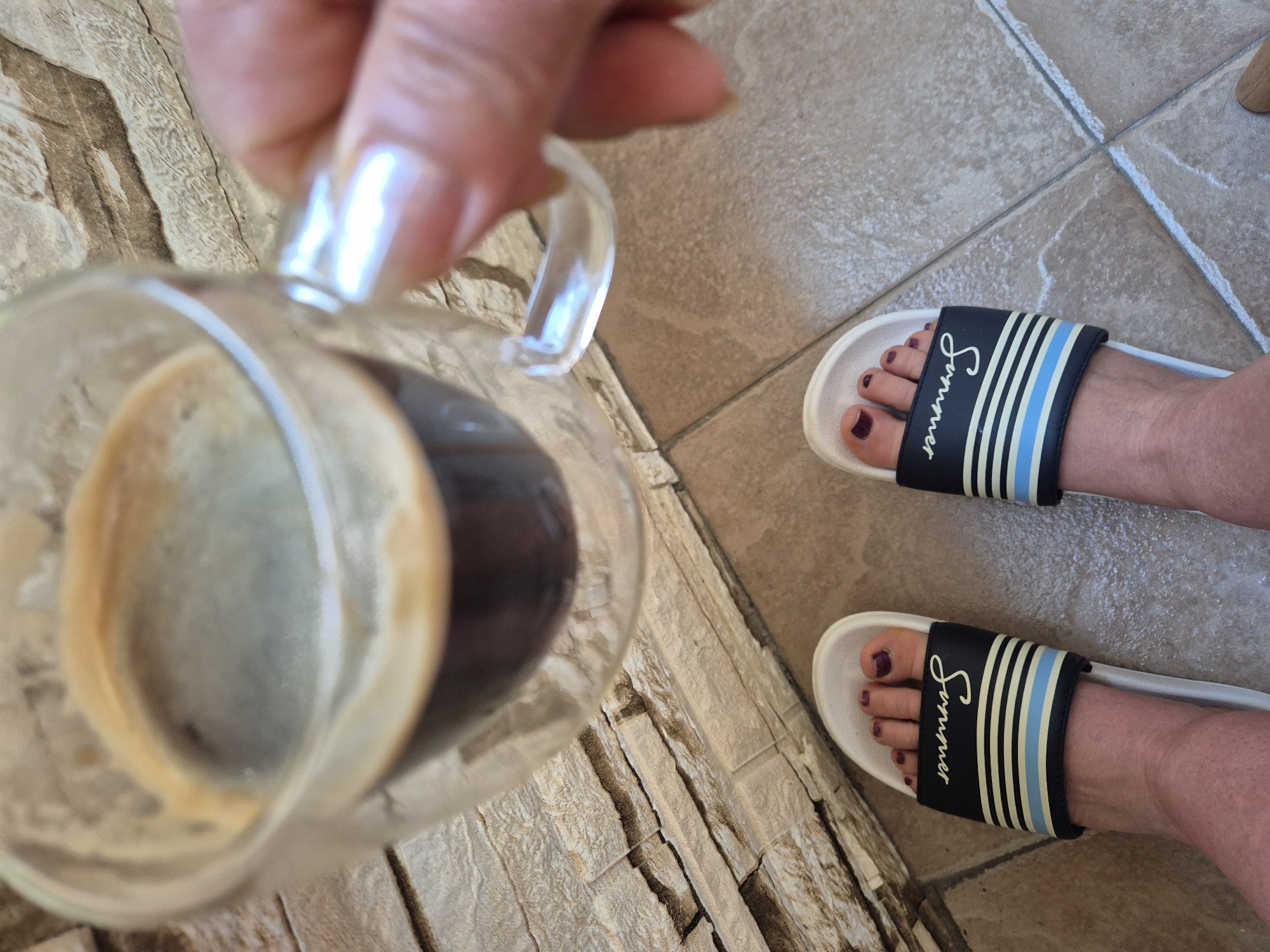 A person's feet, wearing white slides with black straps and blue and white stripes, are visible on a tiled floor. The slides have "summer" written in cursive on the straps. The toes are painted with dark purple nail polish. The person is holding a clear glass mug filled with dark coffee. The background consists of a stone wall.