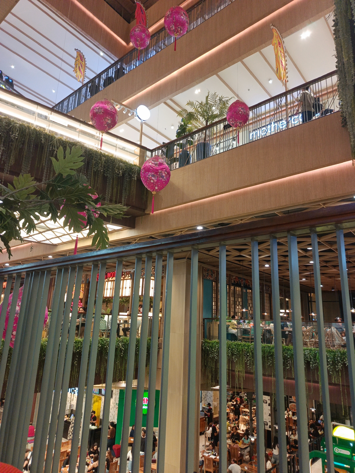 This is a photograph taken inside a multi-level shopping mall. The perspective is from a higher floor, looking down into a central atrium. The mall features a modern design with warm wood tones on the walls and ceilings, accented by soft, recessed lighting. Hanging from the upper levels are several large, pink, spherical lanterns decorated with floral patterns, some with red tassels. Greenery, in the form of hanging plants and potted ferns, is strategically placed throughout the mall, adding a natural element to the interior.
On the various levels, there are balconies and railings overlooking the central space. Several people are visible on these balconies and in the open areas, walking or standing. In the lower levels, restaurants and shops are discernible. Tables and chairs suggest dining areas, and some patrons are seated. The overall atmosphere appears to be one of a bustling commercial space.
The scene strongly suggests that this is a large, modern indoor shopping center with a focus on creating an aesthetically pleasing and inviting environment, likely adorned for a festive occasion given the prominent lanterns. Based on the available information, this location is in Denpasar, Indonesia.