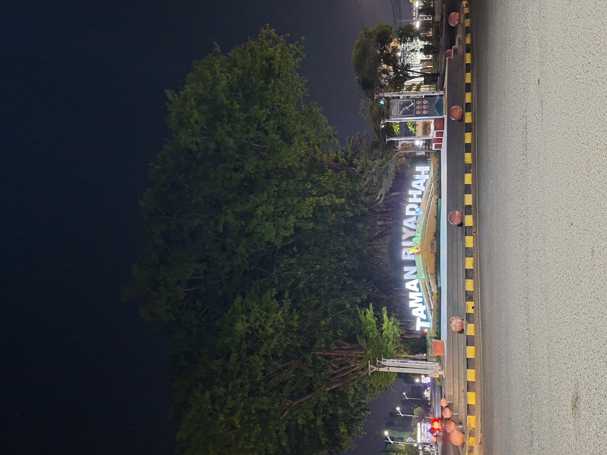 The image displays a nighttime scene of Taman Riyadah in Lhokseumawe, Indonesia. A prominent feature is a large, brightly illuminated sign spelling "TAMAN RIYADHAH" in white capital letters. The sign stands on a landscaped base that appears to be lit with green lights.
To the left of the sign, the scene is dominated by numerous large, dark green trees with dense foliage, indicating a park or green space. The trees extend high into the dark, clear night sky.
On the right side of the image, an asphalt road is visible, bordered by a curb featuring distinct black and yellow stripes. Several spherical, reddish-orange bollards are spaced along this curb. In the background, numerous streetlights emit a bright, artificial glow, illuminating the road and distant areas. Blurry red lights in the far distance suggest the presence of vehicles, possibly moving along the street.
A small green traffic light is visible behind the main sign, along with another structure bearing icons, which might be an informational panel for the park. The overall setting is an urban park entrance or public area beside a road, well-lit for nighttime visibility. No specific individuals or discernible activities are captured, but the illuminated infrastructure suggests a functional and accessible public space. The weather appears clear and calm.