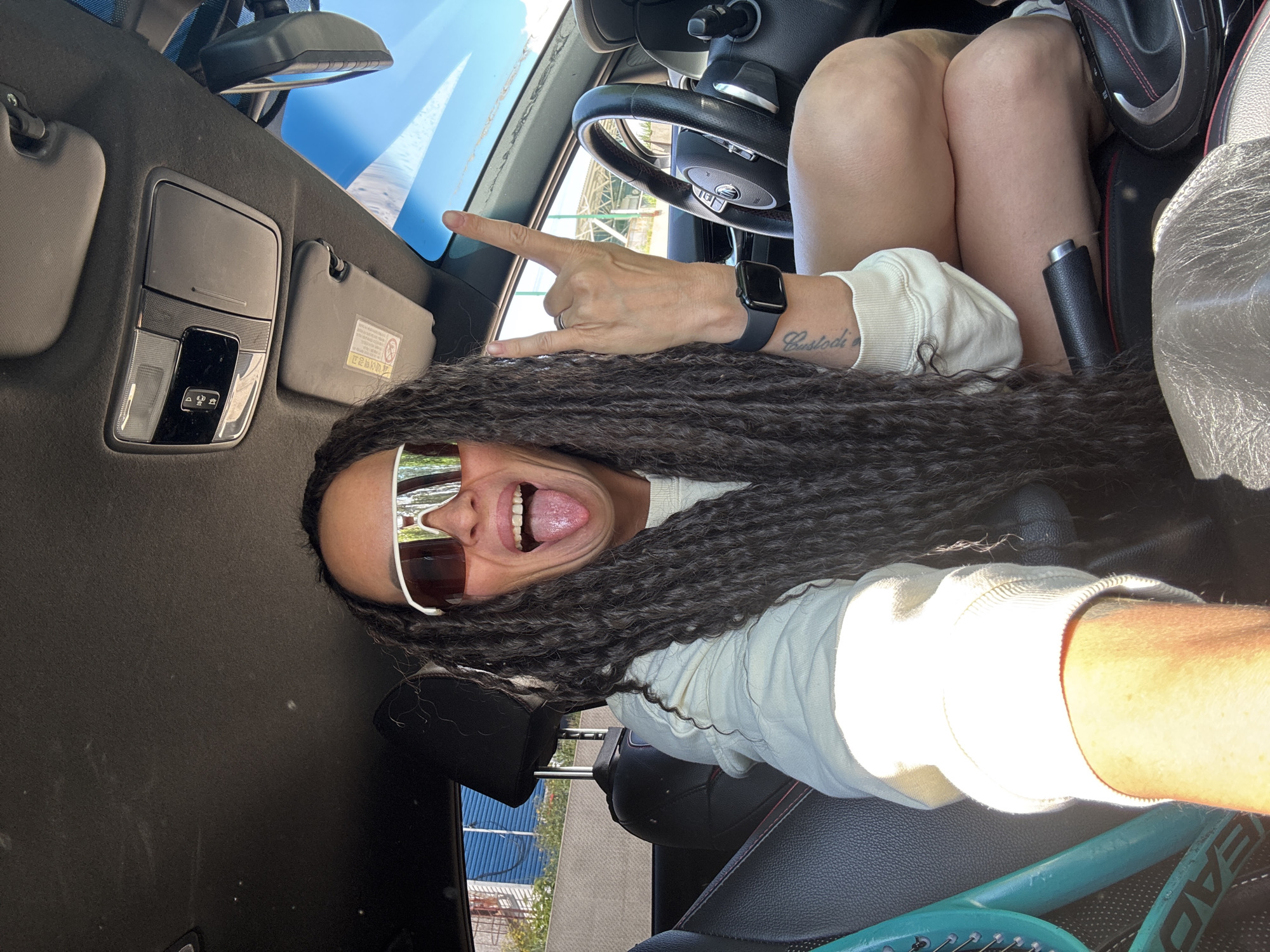 This vibrant, high-angle selfie captures an individual inside a car, likely in Chaikovsky, Russia, on a bright, sunny day. The subject is an adult with long, dark, braided hair, wearing sunglasses and a light-colored long-sleeved top. They are smiling widely with their tongue playfully extended and making a "rock on" hand gesture with their left hand. An Apple Watch is visible on their left wrist, and a tattoo, partially reading "Custodi," adorns their forearm.
The car's interior is detailed, showcasing a dark-colored overhead console, a rearview mirror, and sun visors, one of which displays an illegible warning label. A black leather steering wheel and car seats, possibly with red accents, are also visible. Through the windshield, a clear blue sky and blurred glimpses of the outside environment, including greenery and what appear to be distant structures, are seen. A teal-colored tennis racket with "HEAD" branding is partially visible in the lower right, suggesting a recreational context. The overall impression is one of playfulness, joy, and energy.