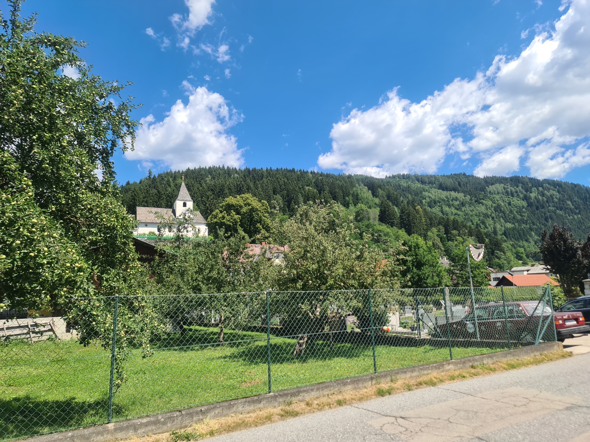 A sunny day scene depicts a suburban or rural area with a prominent church spire in the background, nestled against a densely forested mountain. The foreground is dominated by a green chain-link fence enclosing a manicured lawn with several trees, some bearing fruit. Beyond the fence, a road is visible, with a section of cracked asphalt. A few parked cars are present, one with a white circular sticker on the rear window. The sky is a vibrant blue with scattered white clouds. The overall impression is one of a peaceful, idyllic landscape. This scene is likely located in or near the city of Feldkirchen in Kärnten, Austria, given the architectural style and natural surroundings characteristic of the region.