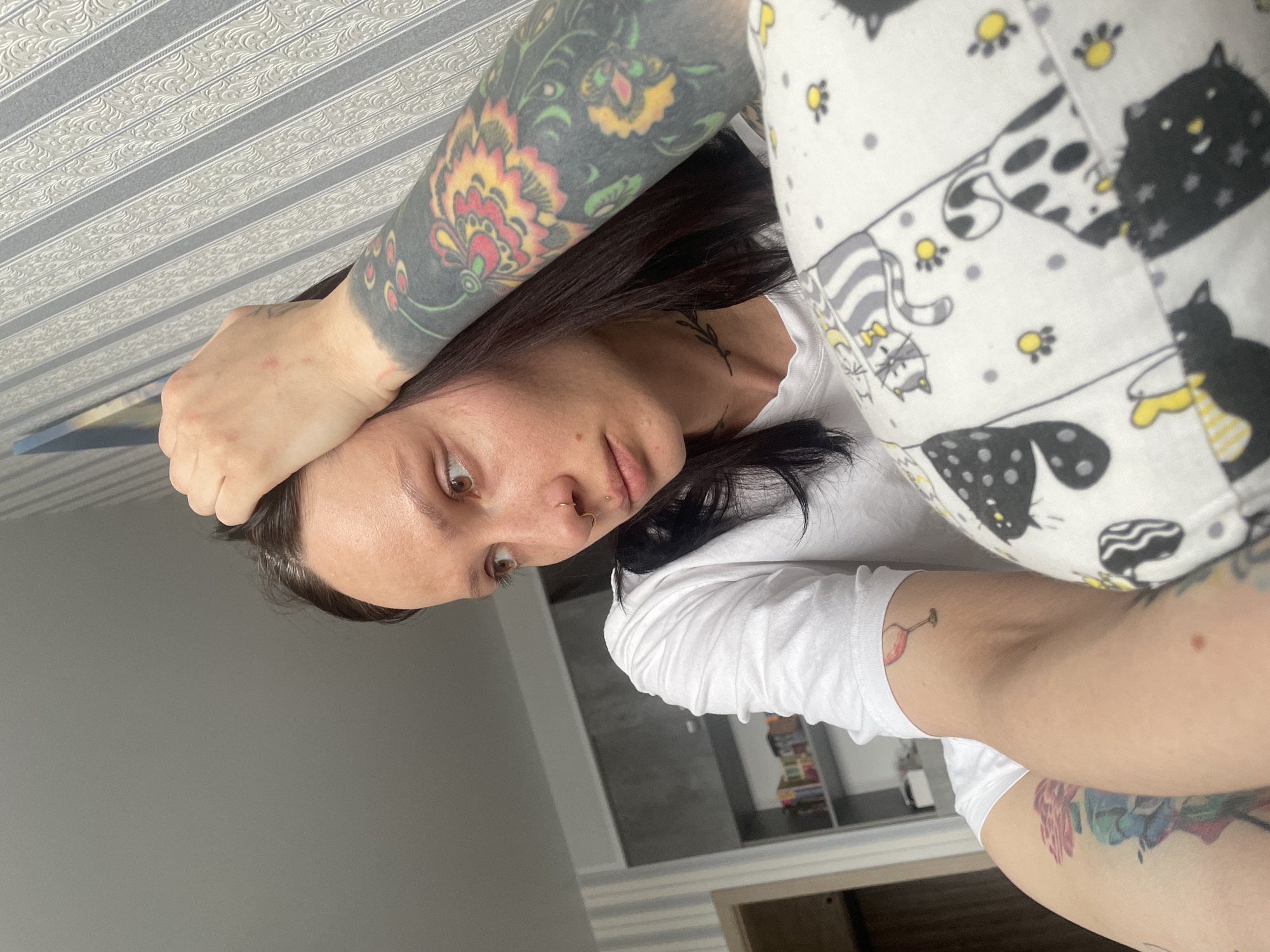 A woman with dark hair and multiple tattoos is taking a selfie indoors. She is wearing a white t-shirt and patterned pajama pants with cat designs. Her left arm is raised and her hand is touching her forehead, while her right arm is extended towards the camera. Her face is tilted down and to the left. The background includes a patterned ceiling, a gray wall, and a glimpse of a room with a shelf. The image was likely taken in a residential space, possibly in Izhevsk, Russia, although this cannot be confirmed definitively from the image alone.