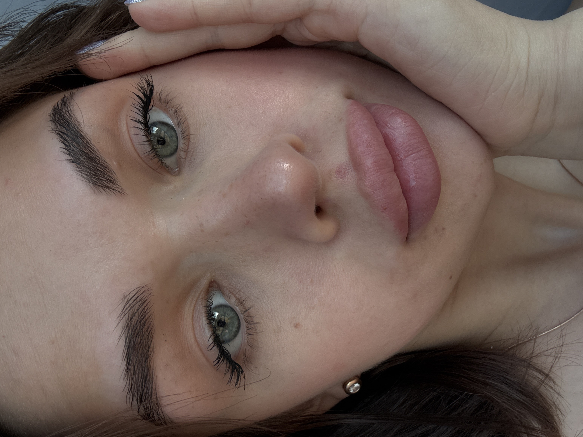 A close-up, angled shot features a woman's face. The background is blurred and indistinct. Her eyes are green and heavily made-up with mascara. The eyebrows are dark and thick. She has fair skin with some freckles. Her lips are full and pink. Her left hand is near her forehead. A small earring is visible on her left earlobe. A thin necklace is also seen near her neck. The location is not discernible from the image.