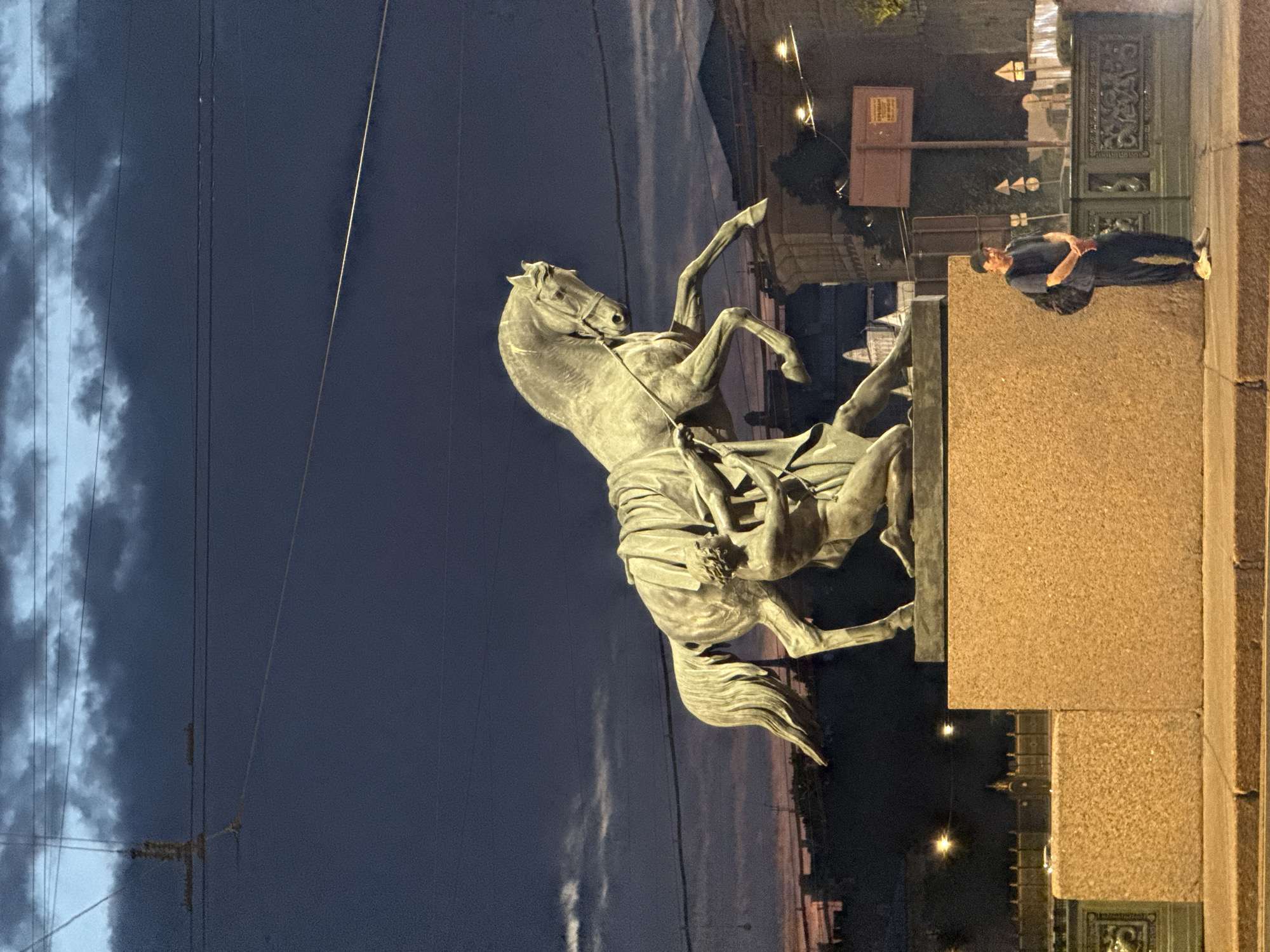 A bronze statue of a man wrestling a horse stands on a granite pedestal. The horse is rearing up on its hind legs, while the man struggles to control it. The man is naked and muscular, with his body twisted as he tries to subdue the powerful animal. The statue is set against a dark blue twilight sky with some clouds visible. In the foreground, a man in a dark t-shirt, dark pants, and a baseball cap stands with his arms crossed, wearing a backpack. He is facing the camera and appears to be looking at the statue. The scene is illuminated by streetlights, casting a warm glow on the man and the pedestal. The background shows buildings and streetlights of the city of Saint Petersburg. This is likely the Anichkov Bridge, known for its four equestrian statues, the "Horse Tamers" by Peter Clodt von Jürgensburg.