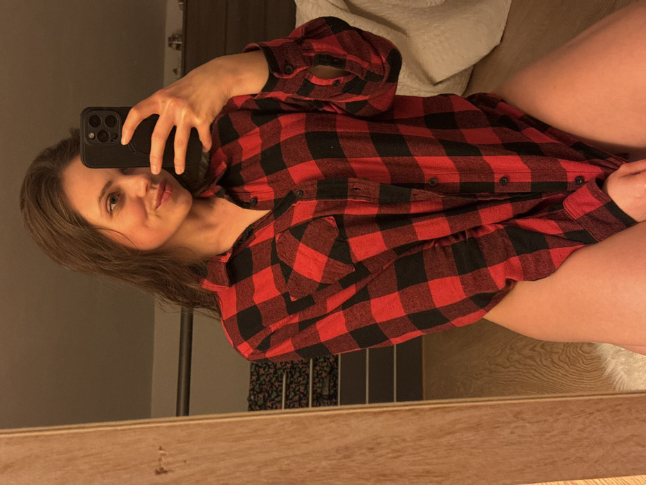 A female subject is captured in an indoor mirror selfie. She has long, dark hair and wears an oversized red and black buffalo plaid flannel shirt, unbuttoned towards the bottom, revealing bare skin on her thighs. She holds a dark-colored smartphone, equipped with a multi-lens camera system, in her right hand to photograph her reflection. The subject displays a slight smile. The environment appears to be a dimly lit private room, consistent with a bedroom. Visible elements include a wooden frame or furniture piece to the left, and in the background, a dark headboard or similar furniture, a white textured textile (likely a blanket or pillow), and a patterned fabric. A white, fluffy textile object is partially visible beneath the subject. There are no identifiable landmarks or geographical features within the image to determine a specific location in the city of Cava de' Tirreni, Italy.