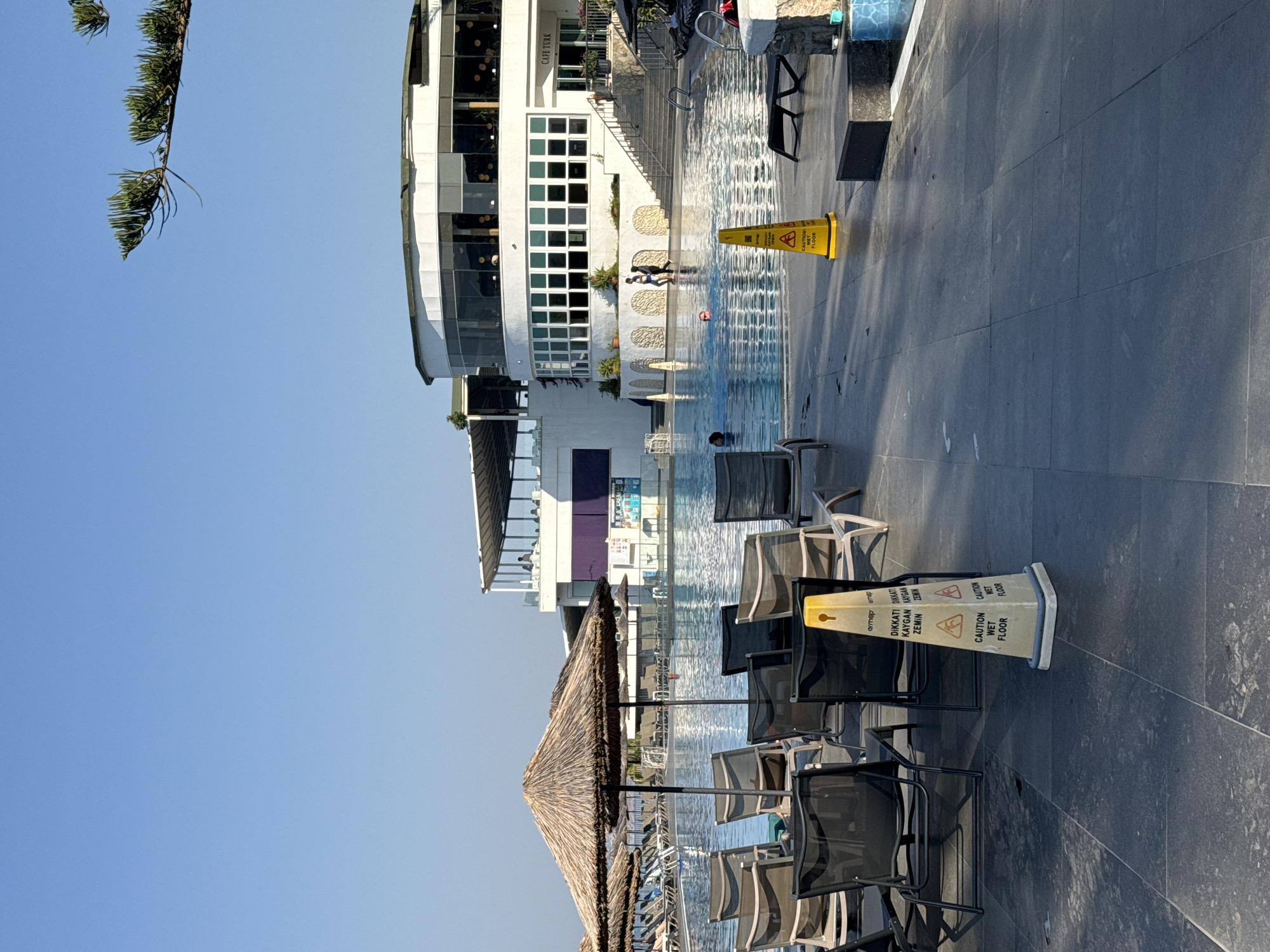 The image depicts a resort pool area in Bodrum, Turkey. In the foreground, several dark-colored lounge chairs are arranged on a dark stone patio. Two yellow caution cones with "CAUTION WET FLOOR" and Turkish text are present on the patio. A large swimming pool with clear blue water occupies the midground, with several people swimming and walking in and around it. Straw umbrellas are strategically placed around the pool area. In the background, a modern white building with large windows and a curved facade houses a restaurant or cafe, with tables and chairs visible inside and on an upper-level terrace. The sky is clear and blue.