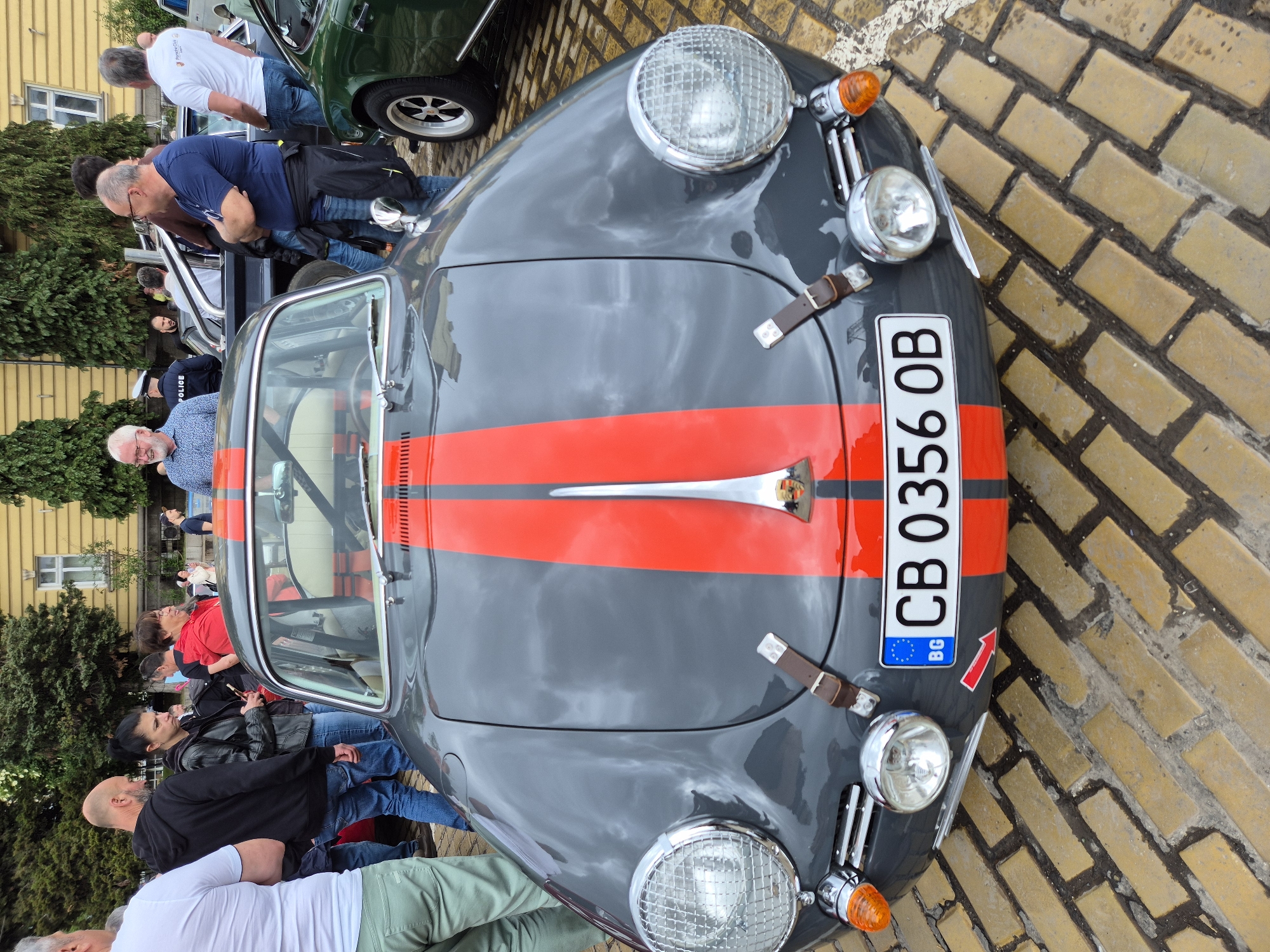 An aerial view captures a gray vintage Porsche 356 with a red racing stripe prominently displayed on the hood. The car bears a Bulgarian license plate “CB 0356 OB.” The vehicle is parked in a public space, likely a street or square paved with yellow bricks, a distinctive feature of downtown Sofia, Bulgaria. Numerous people surround the car, suggesting the photo was taken during an event or car show. Some individuals appear to be observing the car, while others are engaged in conversation. The crowd includes a mix of ages and attire, implying a general public audience. Green trees and buildings are visible in the background, contextualizing the urban setting.