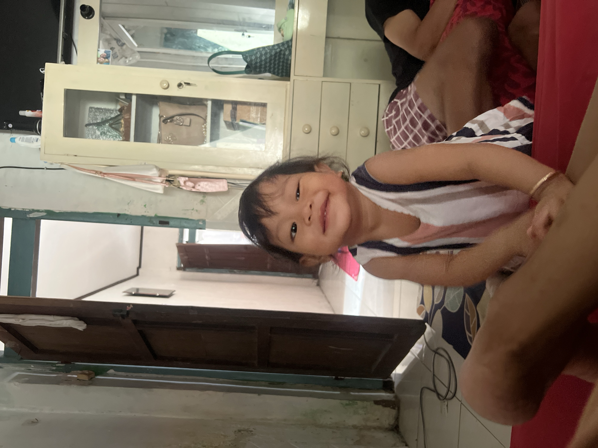 A young child, appearing to be between one and two years old, sits on a red surface, possibly a bed or sofa, in the foreground of the image. The child has dark hair and is looking directly at the camera with a wide smile, showing their teeth. They are wearing a white sleeveless top with vertical stripes of pink, blue, and black. A thin gold bracelet is visible on their left wrist.
Behind the child, to the right, an adult is lying down, mostly obscured by the child. The adult is wearing red and white checkered shorts and a black t-shirt.
The environment appears to be an indoor domestic setting. To the left of the child, a wooden door stands open, revealing a glimpse of a hallway or another room with light-colored walls and a tiled floor. There is a cord on the floor near the door.
In the background, to the right, a cream-colored cabinet with glass doors and drawers is visible. Several items are placed on the shelves within the cabinet, including what appears to be a decorative item resembling the Eiffel Tower on the top shelf, and handbags and other personal items on the lower shelves. A green patterned bag and light green footwear are also visible on a counter surface next to the cabinet.
The walls in the room have a textured and somewhat worn appearance, with some discoloration. The overall lighting suggests it is daytime. The setting is consistent with a home in Kecamatan Pati, Indonesia.