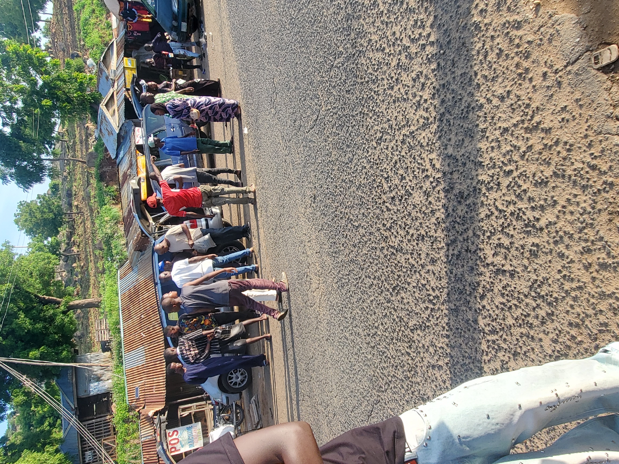 A group of individuals are gathered on a paved road in Ampan, Nigeria. Several vehicles, including a white car, a dark blue SUV, and a dark green sedan, are present. Various structures with corrugated metal roofs are visible in the background, suggesting a market or roadside commercial area. Some people are standing and conversing, while others appear to be observing the surroundings. The ground is asphalt with scattered gravel. The scene is illuminated by natural daylight, casting shadows on the road.