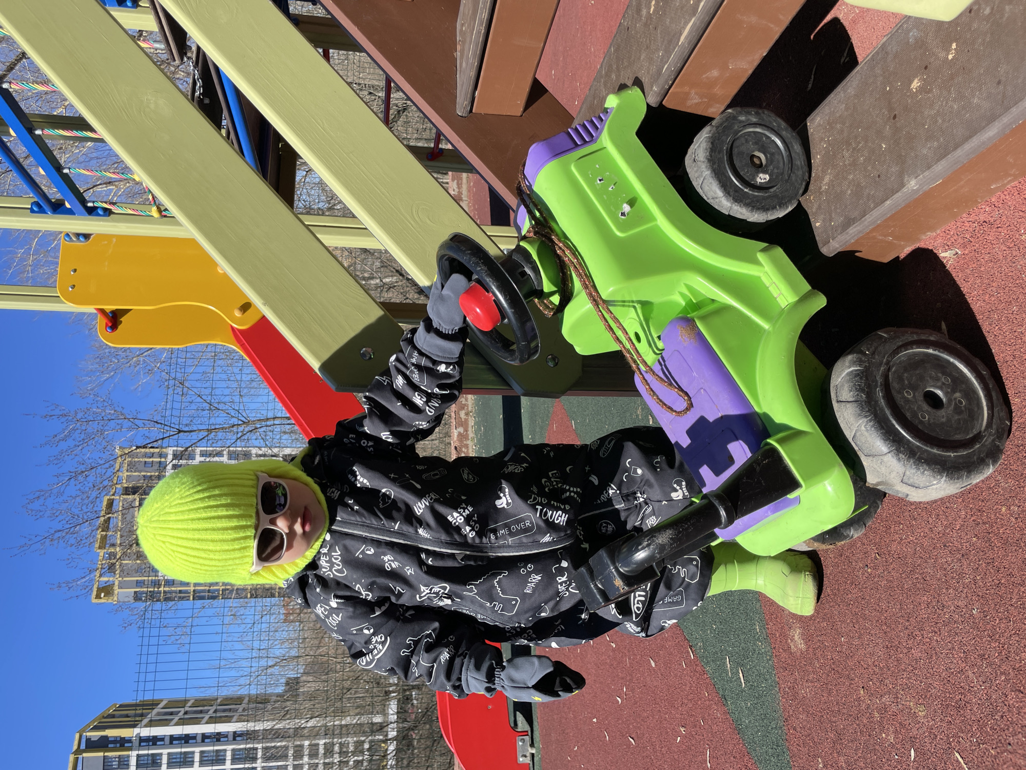 A child in a black and white patterned winter jacket, light green knit hat, sunglasses, and grey gloves is on a playground riding a green and purple plastic toy car. The child is holding the steering wheel of the car. The playground is constructed of wood, plastic and metal, with various sections colored yellow, red, brown, green, and blue. The playground surface is reddish brown. In the background is a clear blue sky with bare trees and a multi-story building with scaffolding. Based on the building style and urban environment, the location is Izhevsk, Russia.