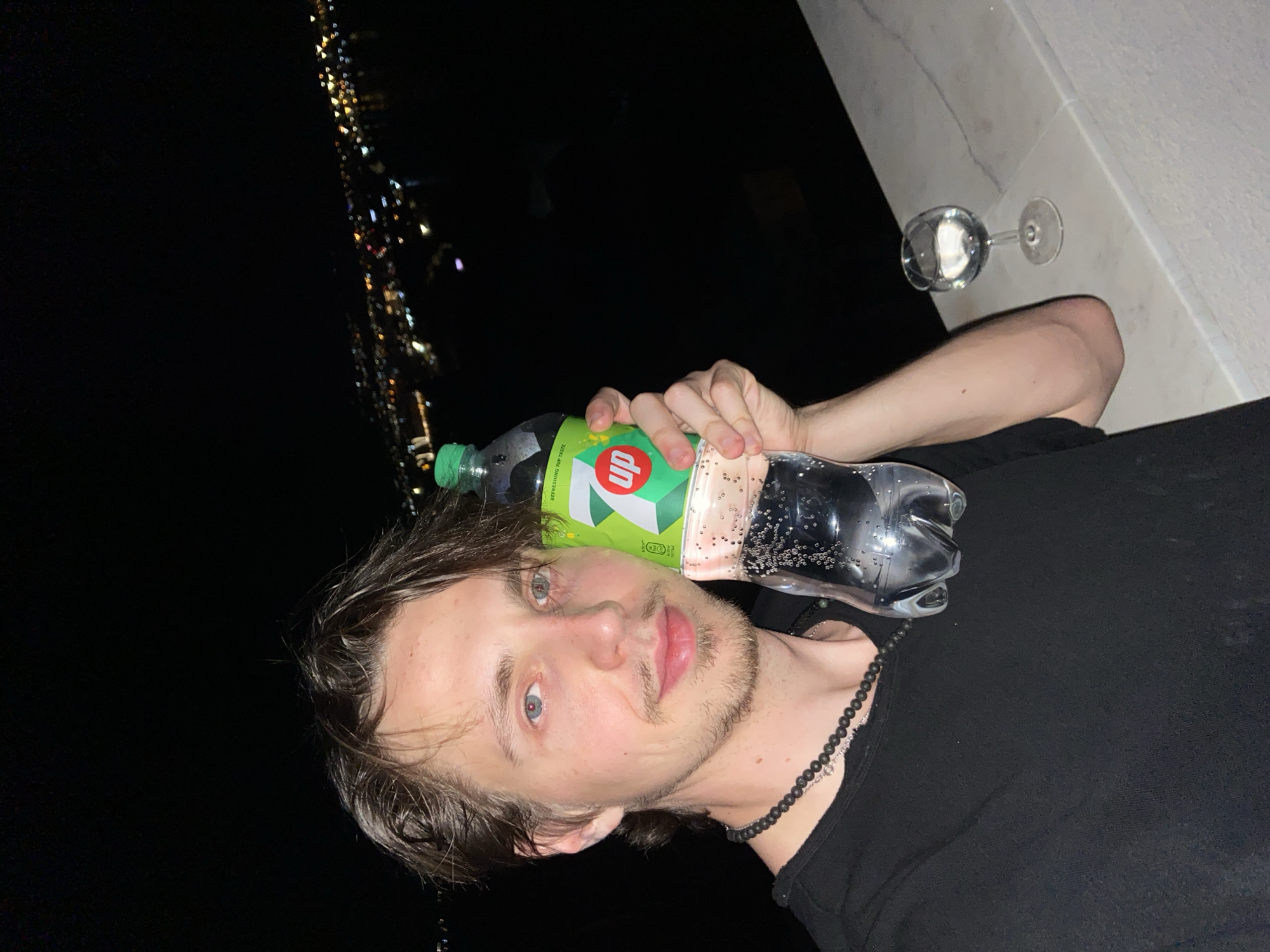 A man is holding a bottle of 7-Up in front of his face. He has light hair and blue eyes. He is wearing a black shirt and a beaded necklace. A wine glass is on a ledge. In the background, lights suggest a city at night. The location is Šušanj, Montenegro.