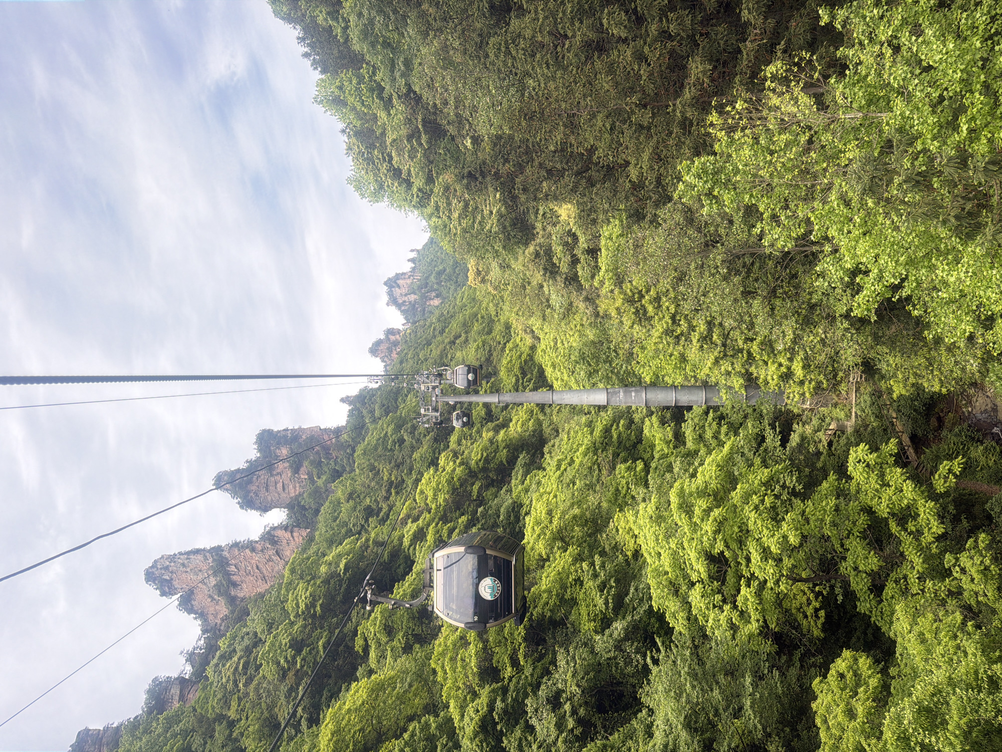 The image depicts a cable car ascending through a lush, mountainous landscape in Wulingyuan, China. The cable car is suspended from thick cables supported by a metal pylon, with several similar pylons visible further up the slope. The mountains are densely covered with trees and vegetation, creating a vivid green environment. In the distance, there are distinct, weathered rock formations. The sky is partially visible, with a mix of clouds and blue patches.