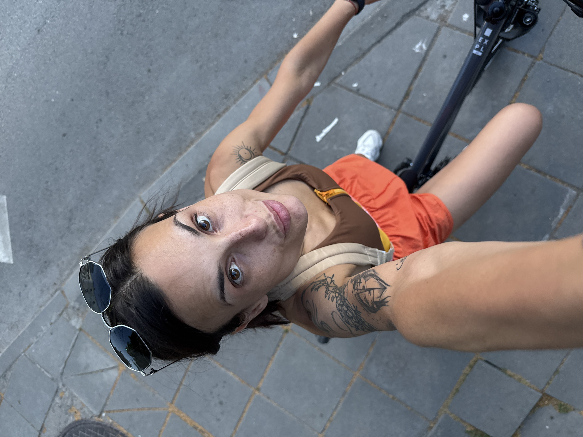 A high-angle, selfie-style photograph shows a woman with dark hair and brown eyes, wearing sunglasses on her head. She is wearing a brown and orange outfit and has tattoos on her arms. She appears to be riding a bicycle or scooter on a paved sidewalk made of gray square tiles. The background consists of more paved sidewalk and a gray street. There are no discernible landmarks that would indicate the city of Pleven, Bulgaria.