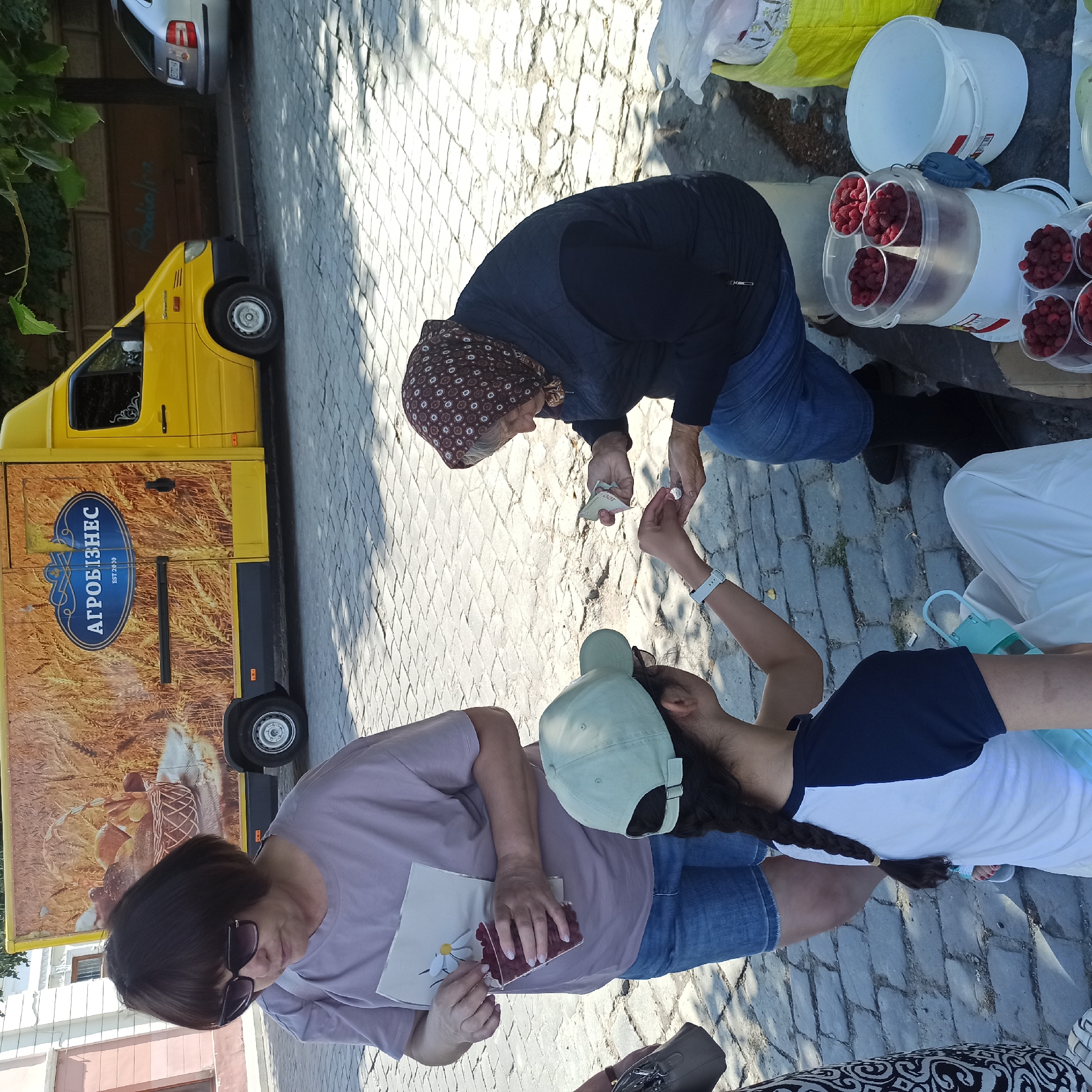 In Chernivtsi, Ukraine, on a cobblestone street, a transaction is taking place between an older woman and a younger person. The older woman, wearing a headscarf and dark clothing, is seated and appears to be handing money to the younger person, who is wearing a light green cap and a t-shirt. The younger person is also wearing a white wristband. Another woman, wearing sunglasses and a light purple t-shirt, stands nearby, holding a clear plastic cup filled with small red berries. In the background, a yellow truck with the logo "АГРОБІЗНЕС" is parked. The scene suggests a street market or informal sale, with baskets of berries visible on a table.
