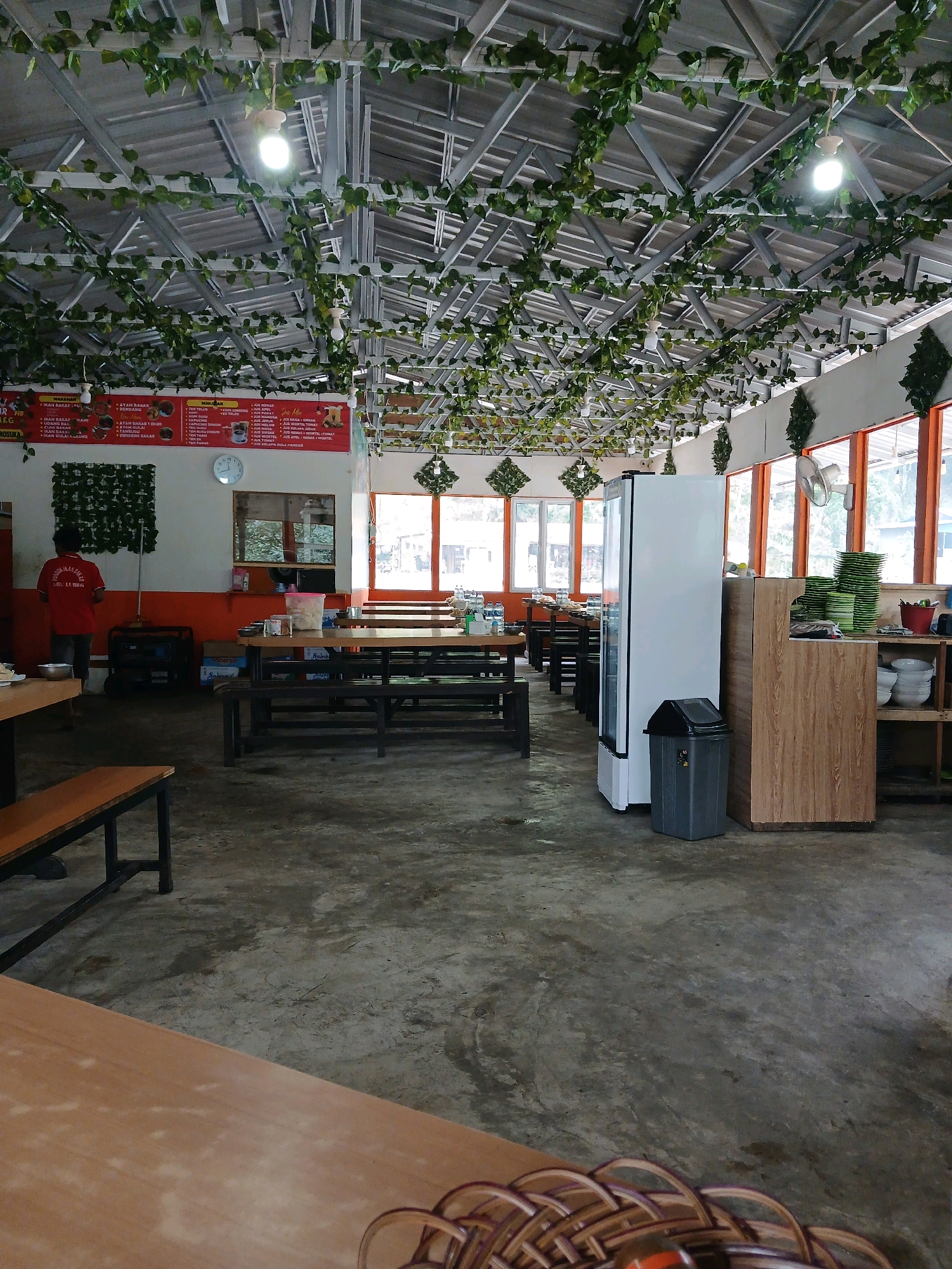The image depicts the interior of a restaurant. The ceiling is adorned with a metal frame and artificial green vines, with several lights suspended from it. In the foreground, a wooden table occupies the bottom portion of the frame. The mid-ground features rows of dark wooden tables and benches, a white refrigerator, a dark gray trash can, and a wooden cabinet with shelves displaying stacks of green plates and bowls. To the left, a person wearing a red shirt is visible near a menu board on the wall. The environment appears to be a casual dining establishment, possibly a food stall or a local eatery. The overall atmosphere suggests an indoor seating area for customers.