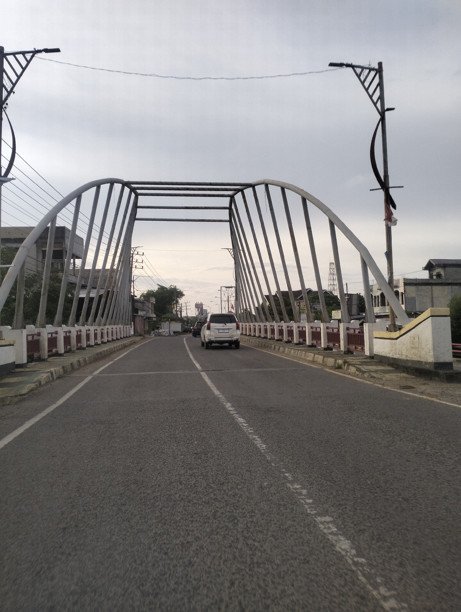 A wide-angle, eye-level shot shows a view of a bridge in Lhokseumawe, Indonesia, on a cloudy day. The bridge has a distinctive arched metal structure with vertical supports and a red and white railing along the sides. The road surface is asphalt with white lane markings. Two white cars are visible on the bridge, one in the foreground and another further ahead. Buildings and trees line the sides of the road beyond the bridge. The sky is overcast and grey. A streetlamp with a zigzag design is positioned on the right side of the bridge.