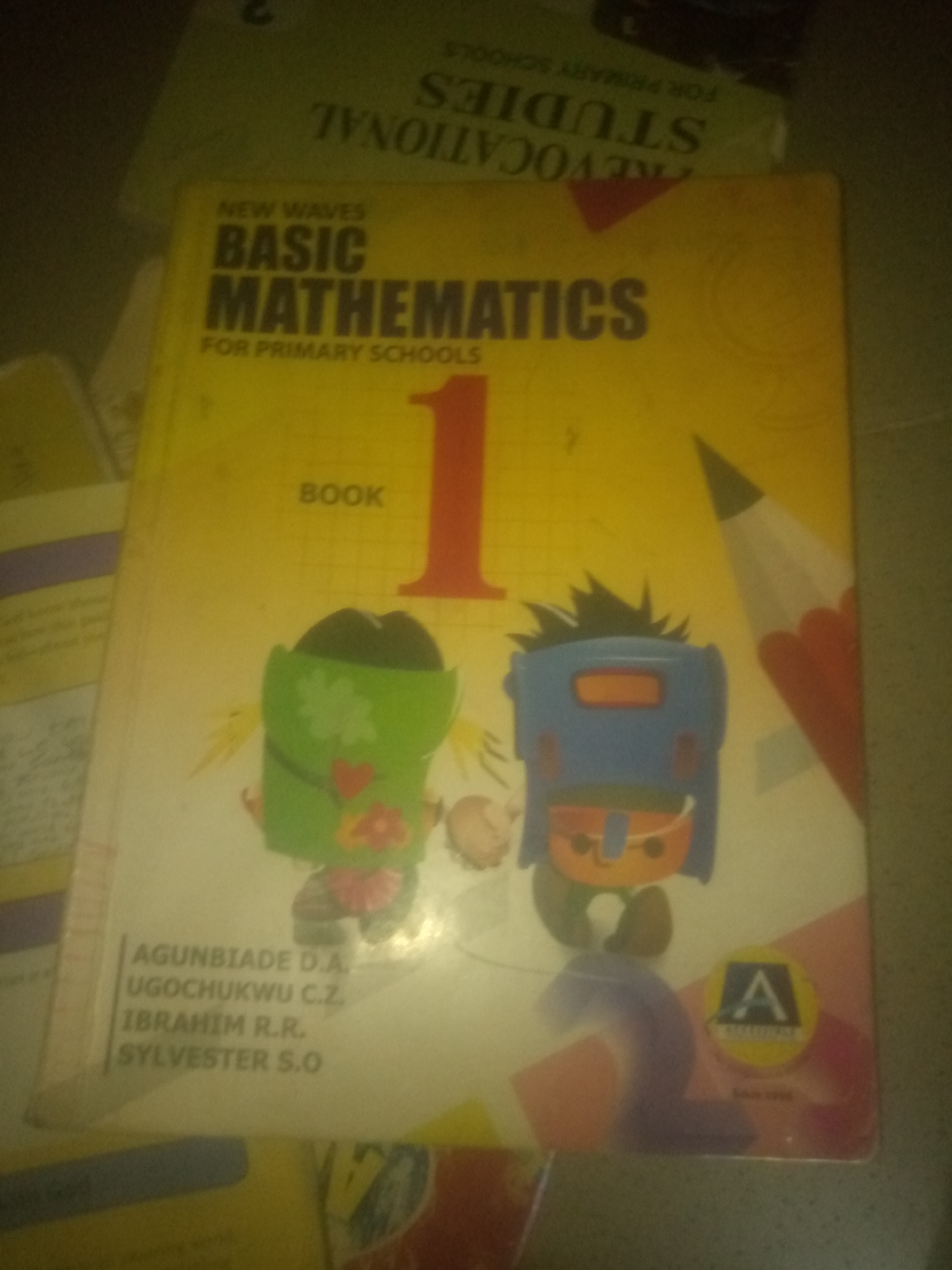 A close-up, high-angle, blurry photograph shows the cover of a yellow textbook titled "NEW WAVES BASIC MATHEMATICS FOR PRIMARY SCHOOLS BOOK 1." The textbook features cartoonish illustrations of two figures with large heads, one green with a red heart and flower, and the other blue and orange with spiky black hair. A large red number "1" is prominent on the cover. Below the title and illustrations, a list of names is visible: AGUNBIADE D.A., UGOCHUKWU C.Z., IBRAHIM R.R., SYLVESTER S.O. In the bottom right corner, a circular logo with the letter "A" is present. The textbook appears to be resting on other books or papers. The lighting is dim, and the image quality is poor, making it difficult to discern fine details of the environment. The image does not provide any information about the location in the city of Jauro Yinu, Nigeria.