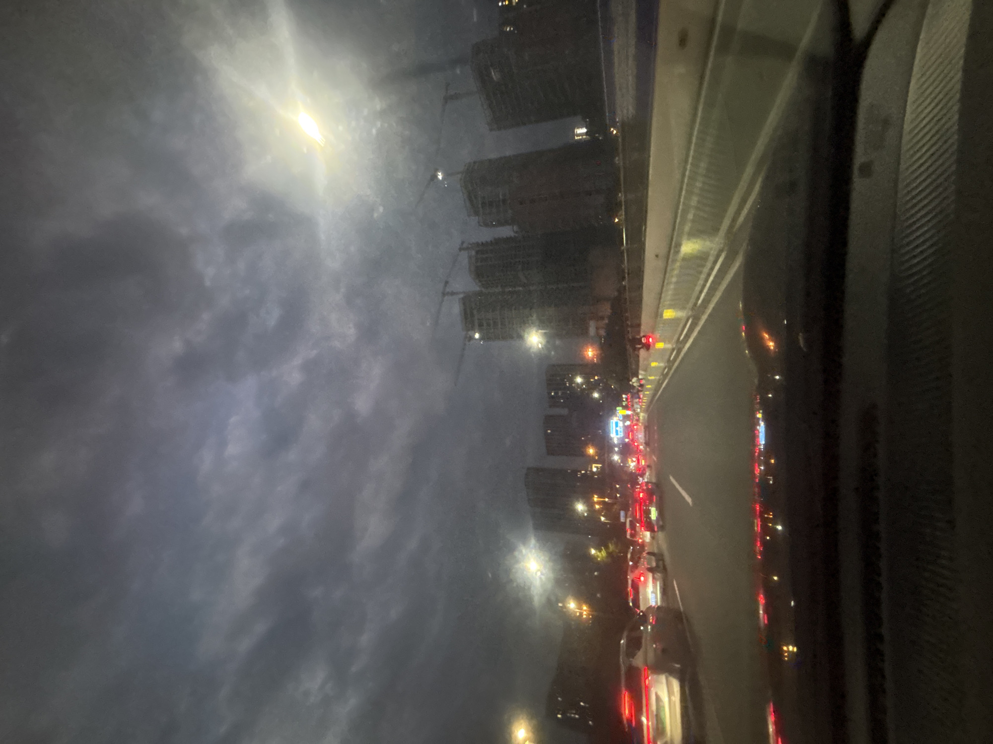The image shows a view from a vehicle traveling on a highway at dusk. The sky is overcast with dark clouds, but a bright light, likely the sun, is breaking through in the upper right portion of the frame. In the distance, several tall buildings, some appearing to be under construction with cranes, are visible. The highway is lined with traffic, with cars visible in multiple lanes, their brake lights illuminated. Streetlights line the road, casting a warm glow. The scene depicts a typical urban commute during twilight hours in Baiyun District, China.